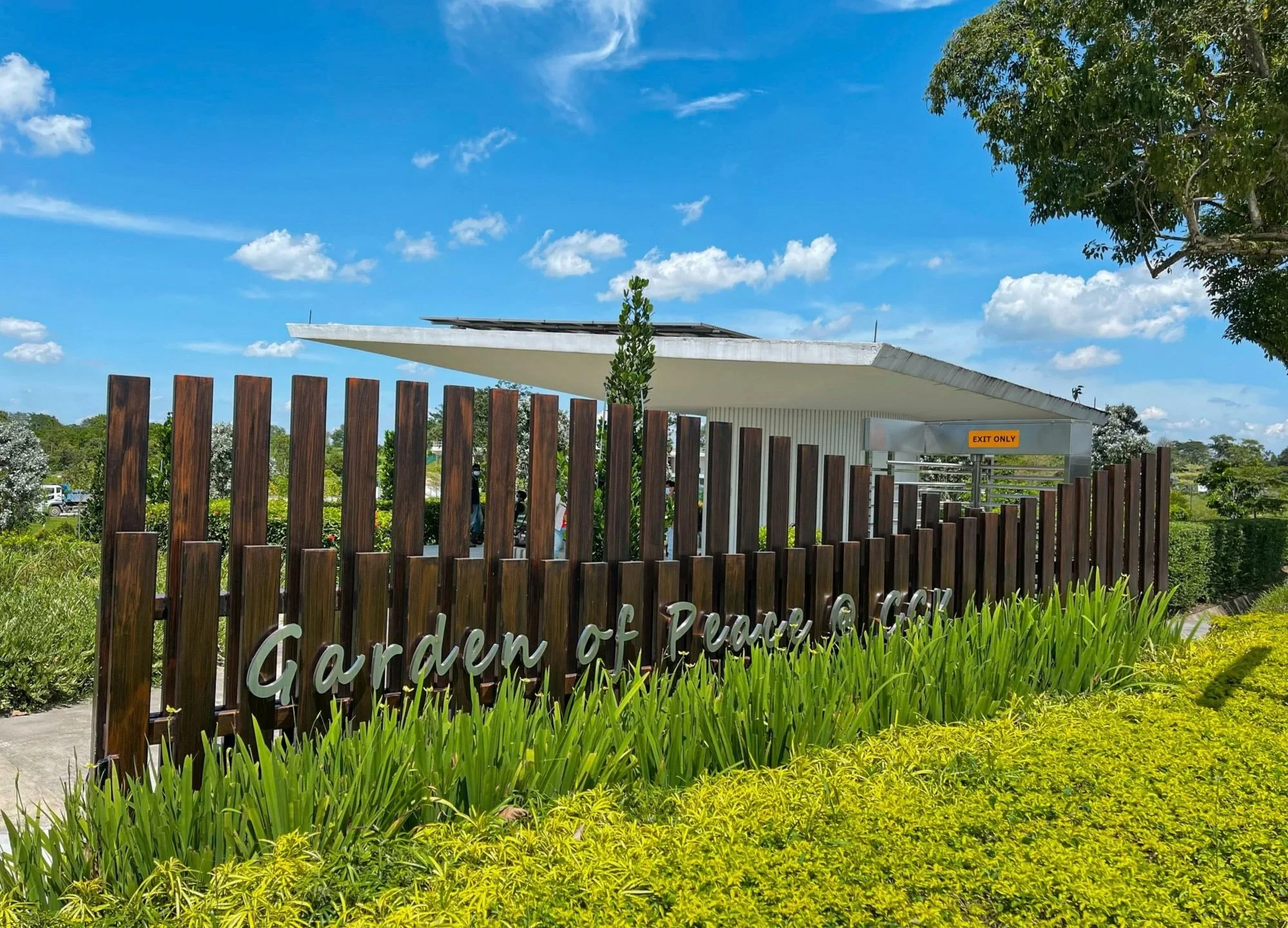 Entrance sign for Garden of Peace, surrounded by greenery, with a modern building and blue sky in the background.