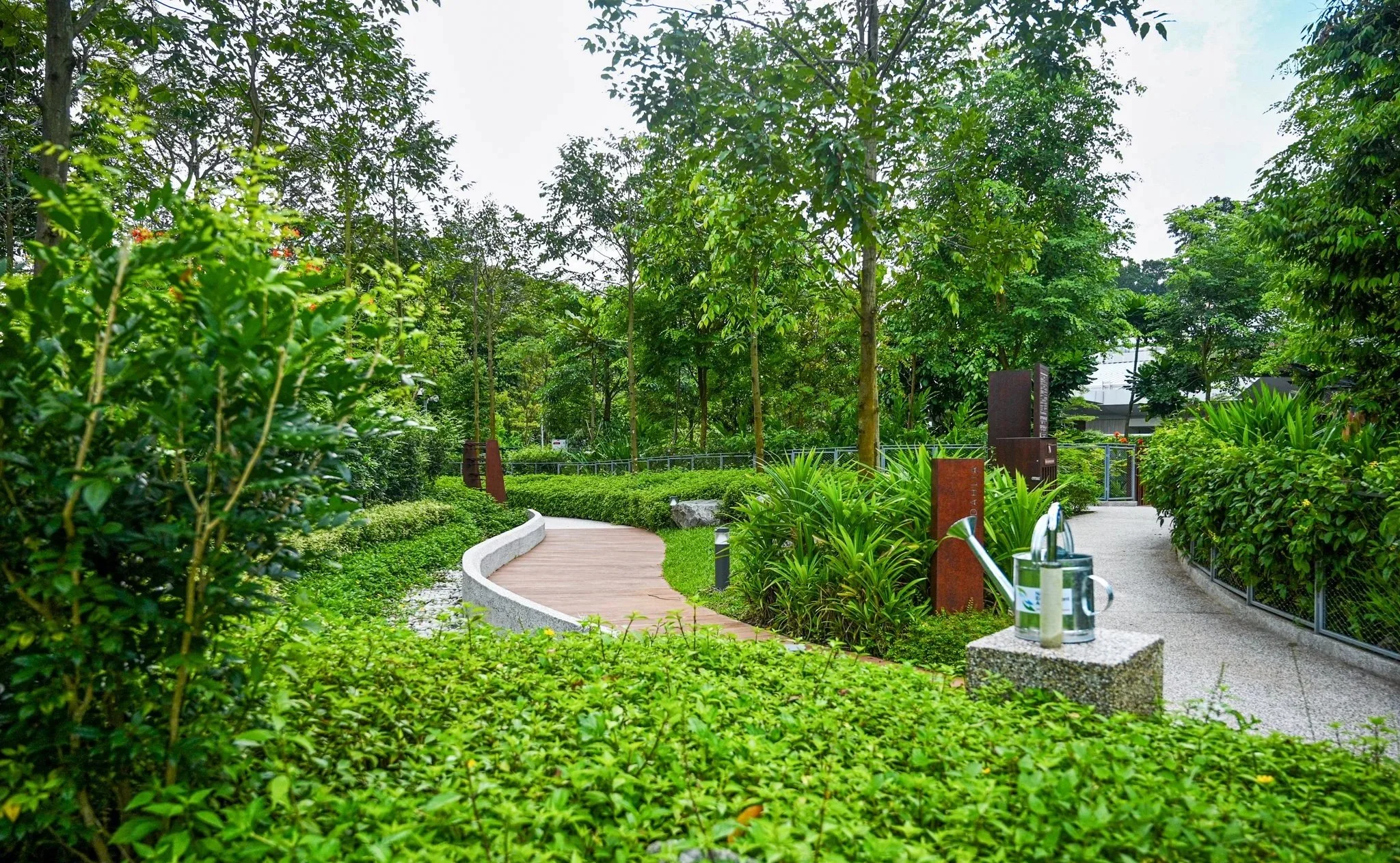 A winding park pathway surrounded by lush green trees and bushes, with a water fountain on a stone pedestal in the foreground.