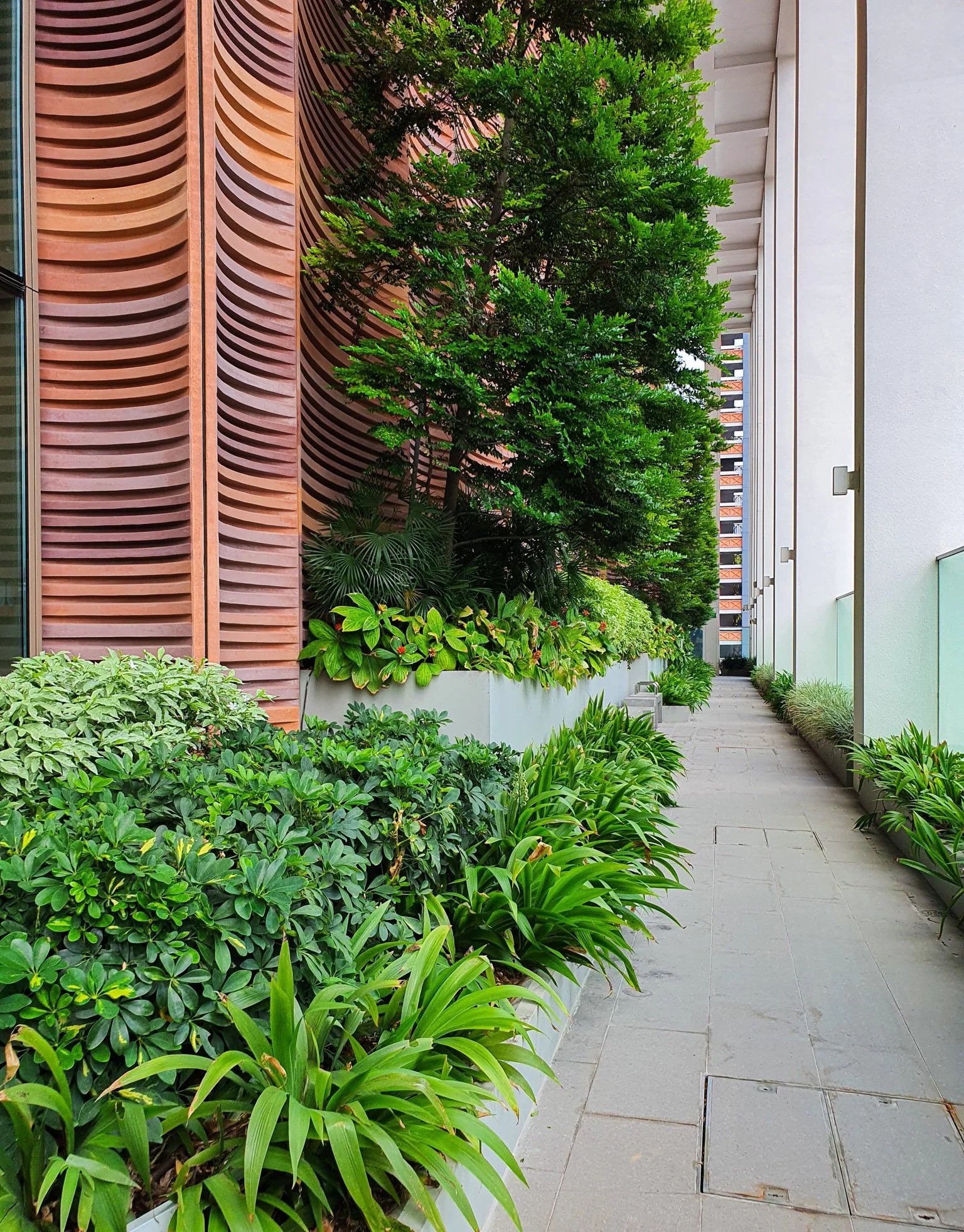 Apartment balcony corridor with lush greenery and potted plants along the walkway, modern building design with patterned wall, and glass railings.