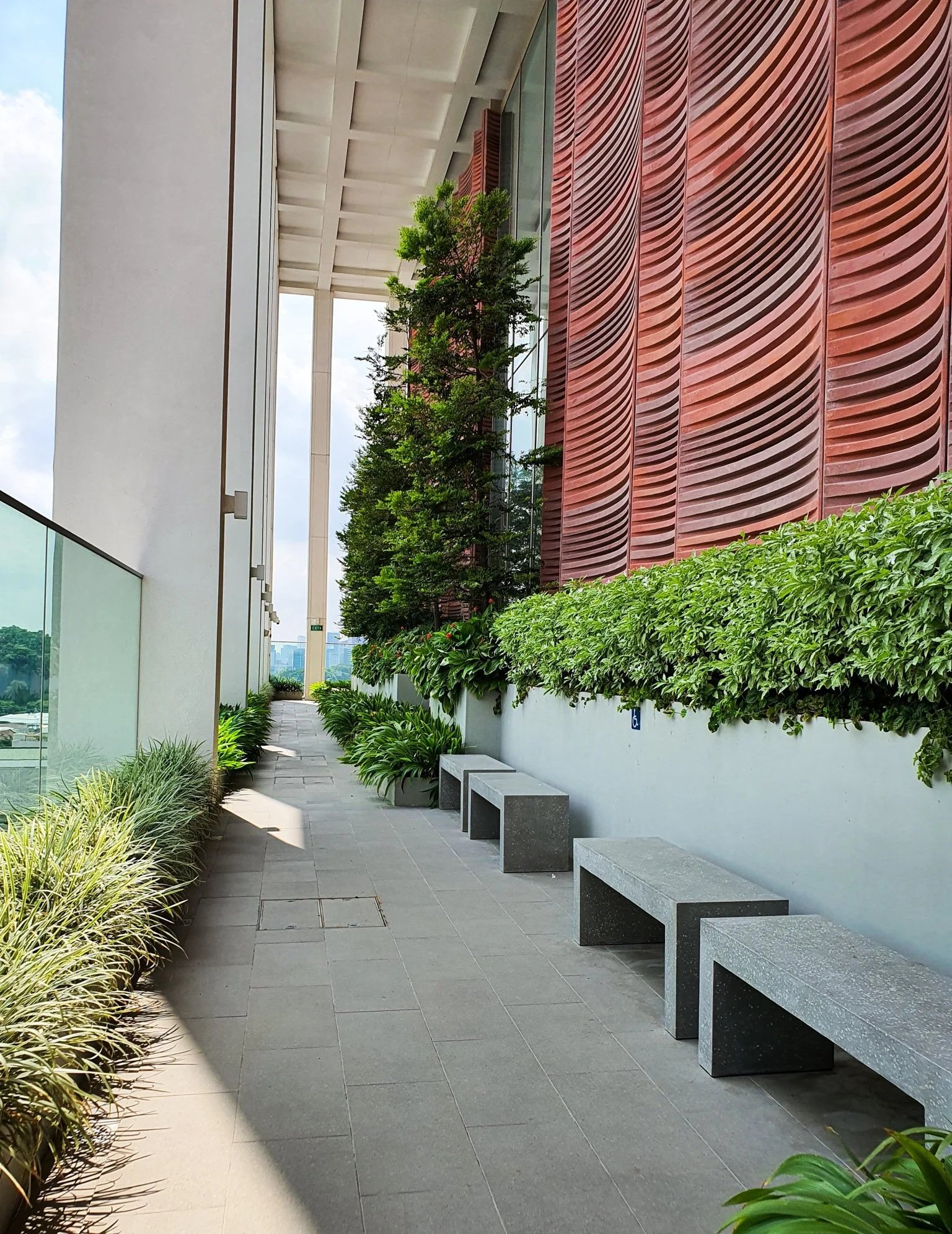 A modern outdoor balcony with concrete benches, green plants, and trees alongside a building with a red textured wall and large glass windows.