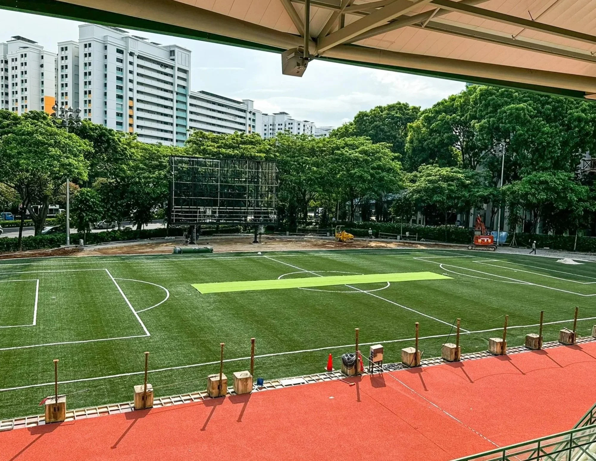 Empty soccer field under construction, with green grass and white lines, surrounded by a red running track. Construction equipment and tools are on the track, with a backdrop of trees and modern white apartment buildings.
