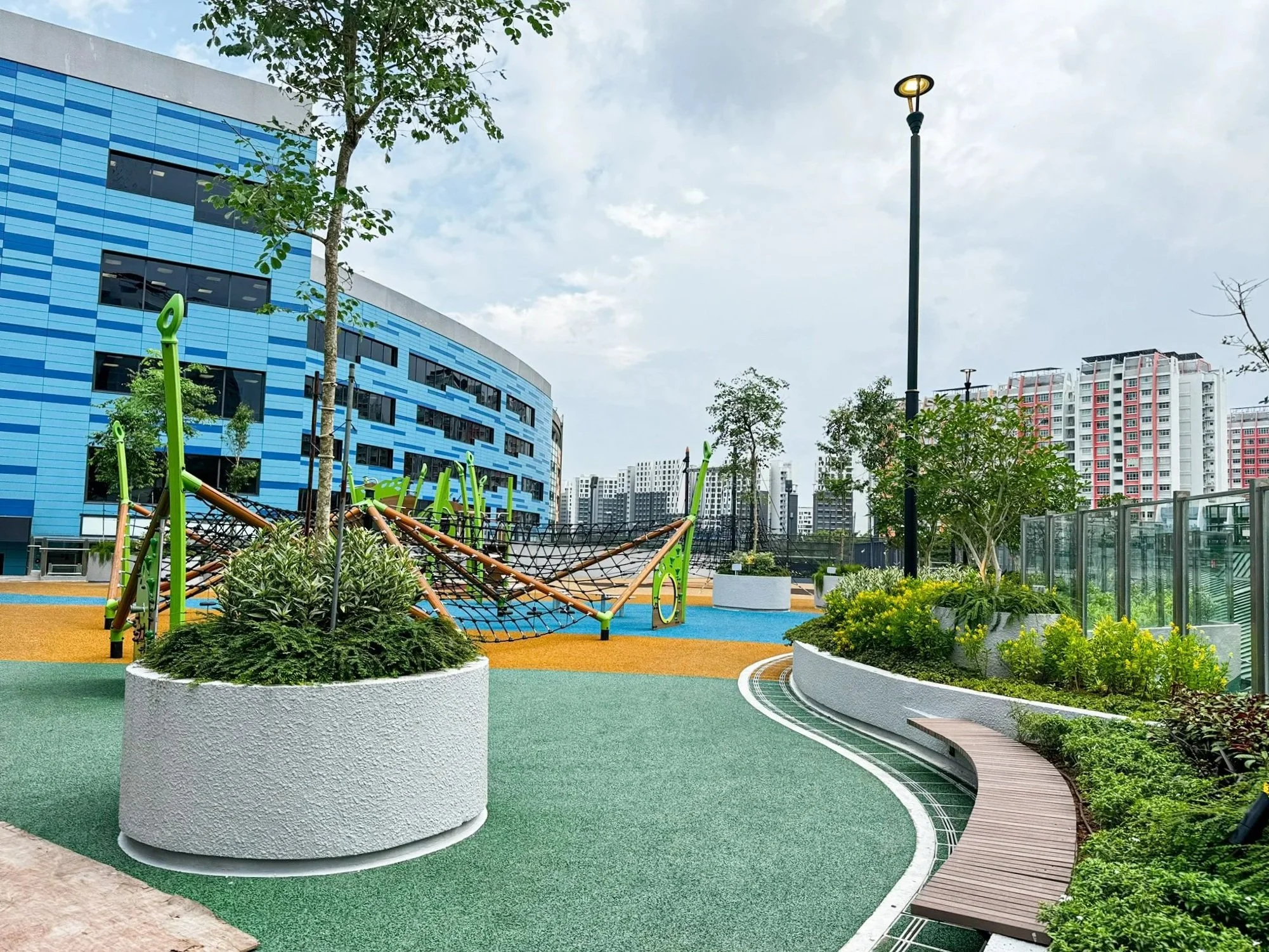 A colorful urban playground with climbing structures and a paved walkway, surrounded by modern high-rise buildings and greenery under a partly cloudy sky.