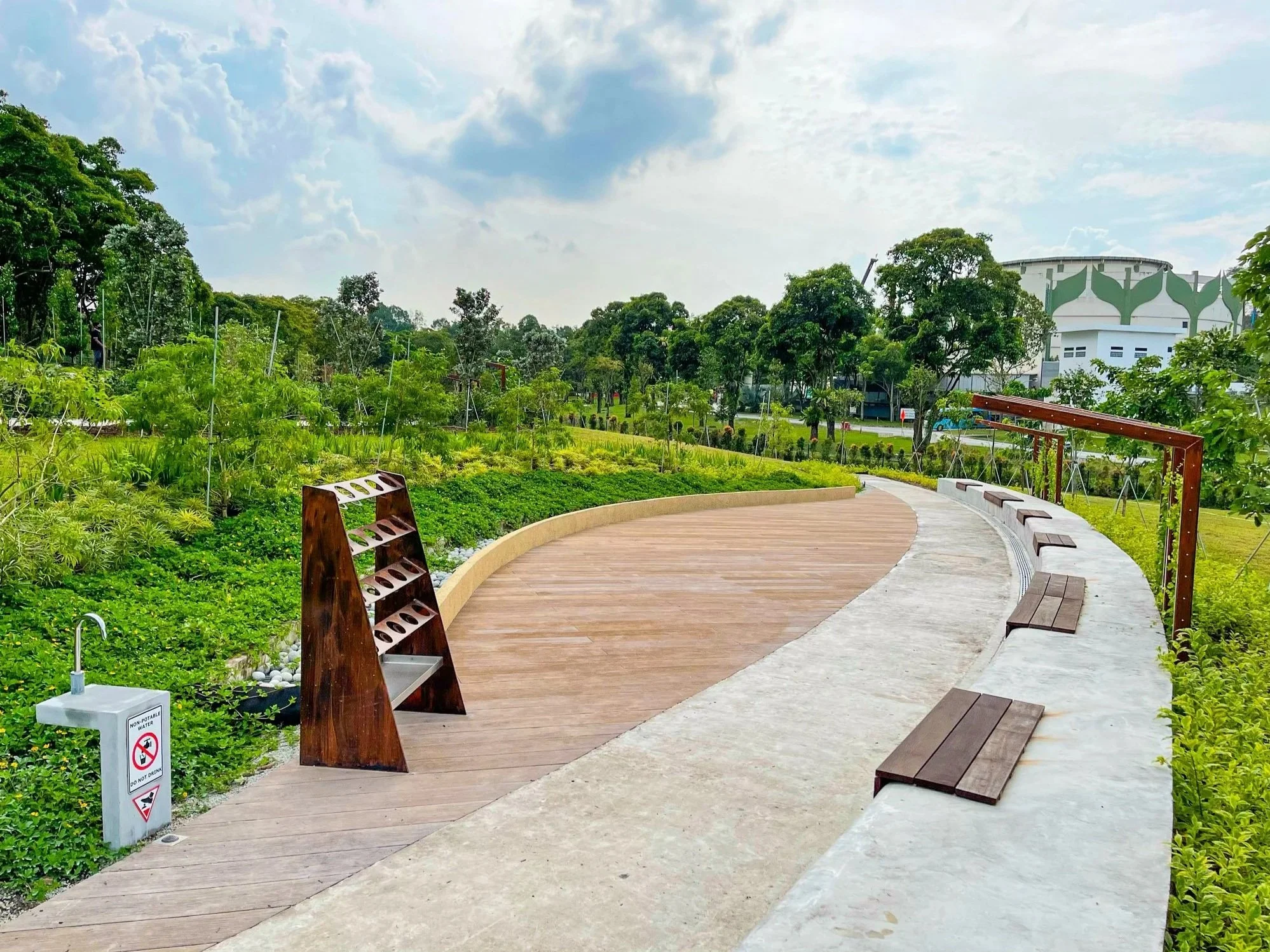 A curved walking path in a park with benches, surrounded by greenery and trees, under a partly cloudy sky.