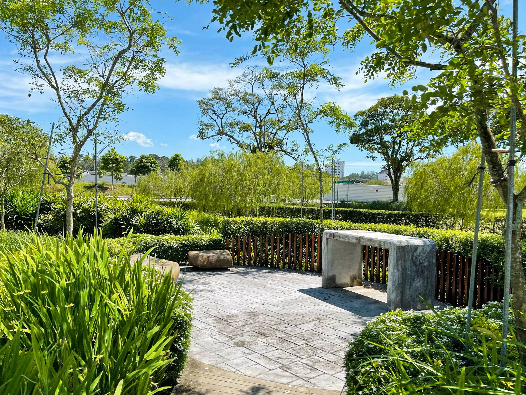 A small park with lush green trees and bushes, a concrete bench, and stone seating. The sky is bright blue with a few clouds, and there are buildings in the background.