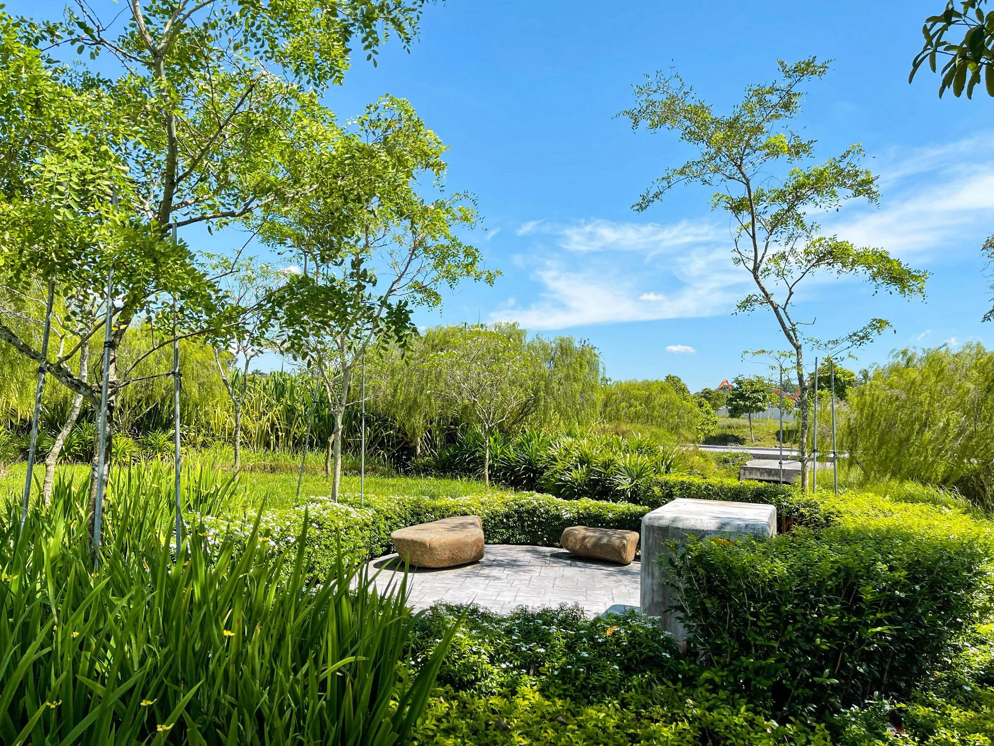 A lush green park with various trees, bushes, and grasses under a blue sky with some clouds. There are large rocks and a paved seating area in the center.
