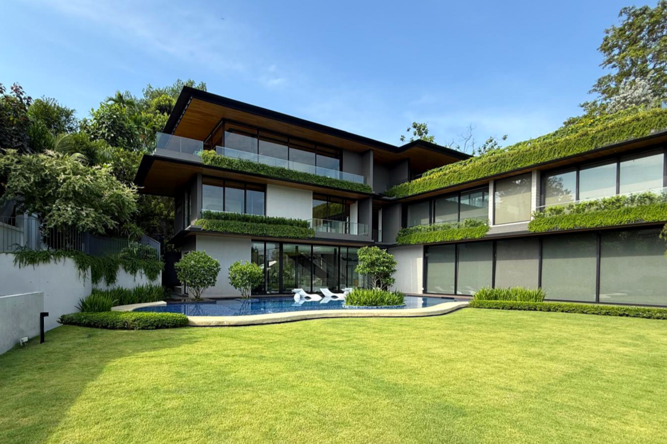 Modern multi-story house with large glass windows, green hedges, trees, a swimming pool, and a well-manicured lawn under a blue sky.