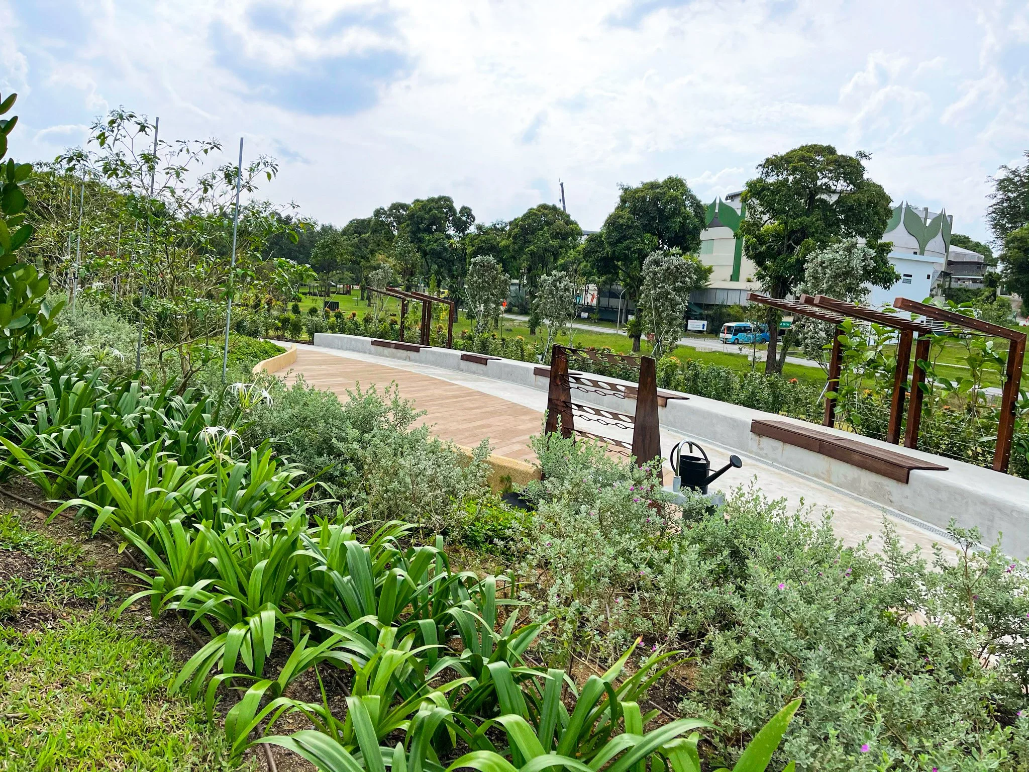 A park with lush green plants, a curved pathway with benches, trees, and a white building in the background under a partly cloudy sky.