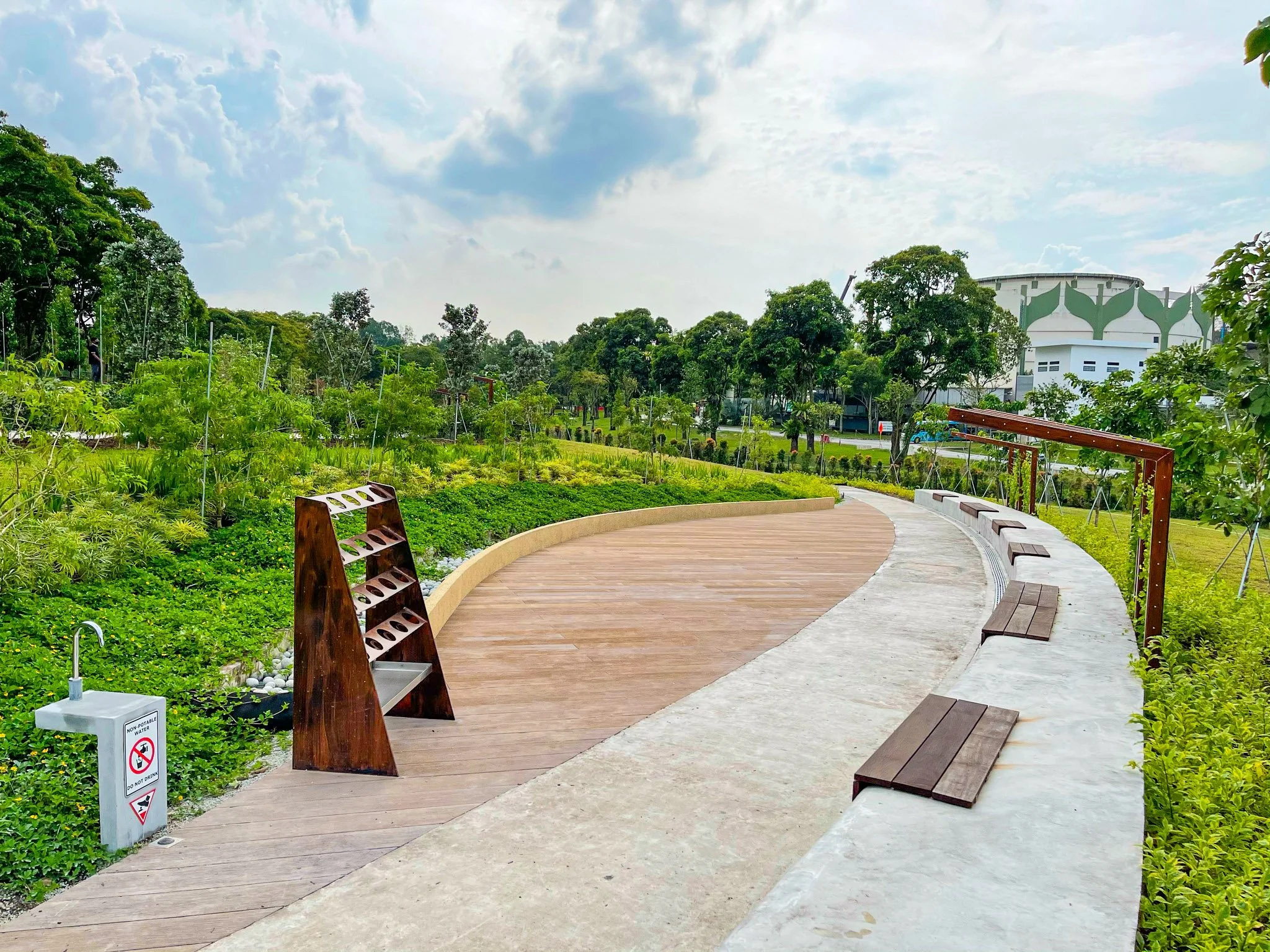 Curved park pathway with benches, greenery, trees, and a modern building in the background under a partly cloudy sky.