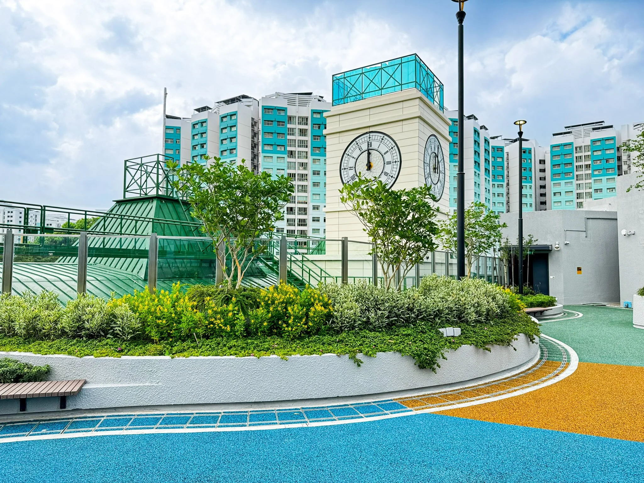 City rooftop with a clock tower, high-rise buildings, green plants, trees, and colorful patterned flooring.