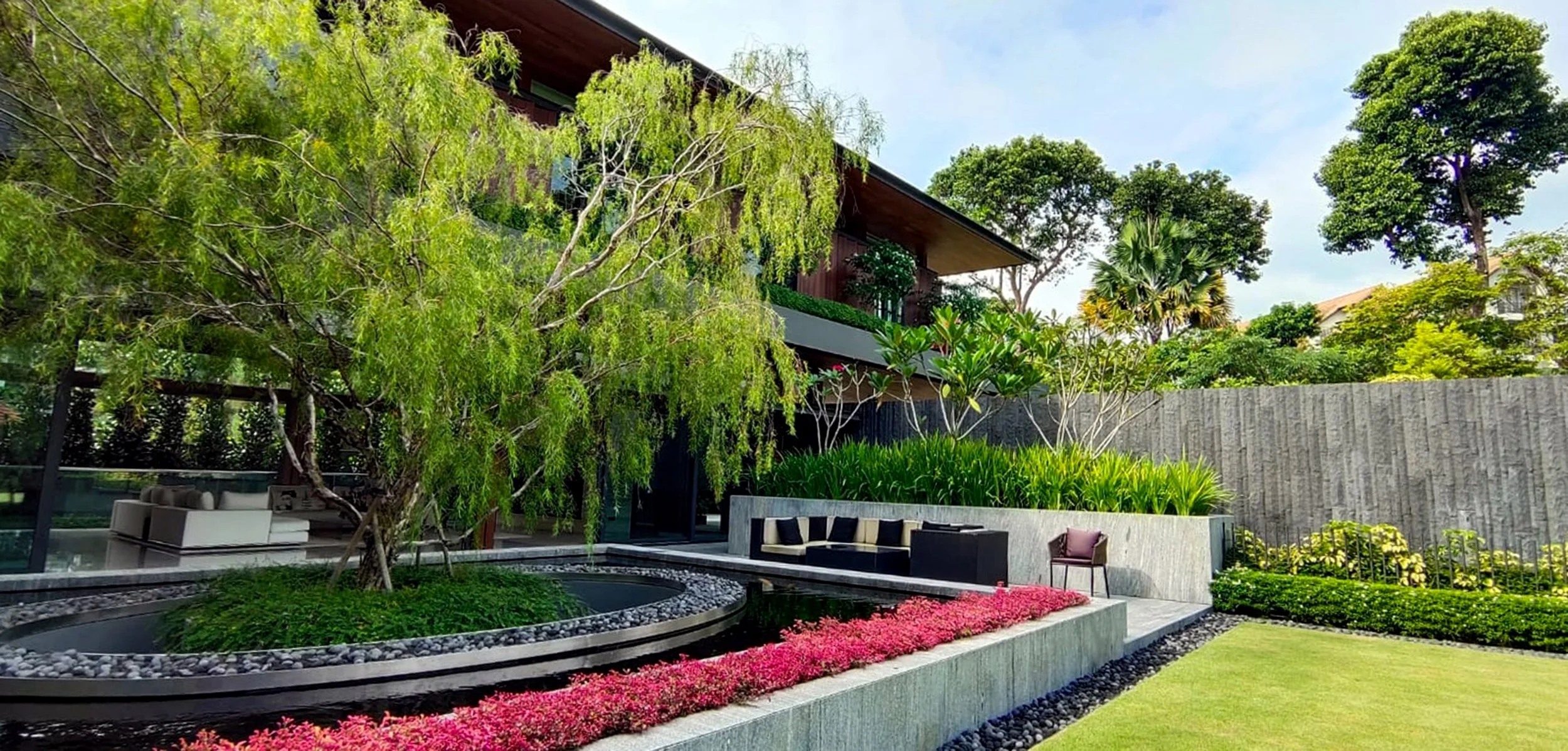 Modern backyard with lush greenery, a small pond, pink flowers, and outdoor seating, with a house and tall trees in the background.