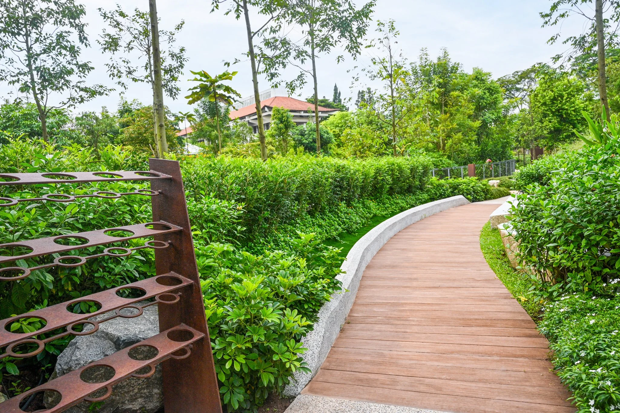 Curving wooden pathway through a lush green garden with various plants and trees, a metal rack on the left side, and a building with a red roof in the background.