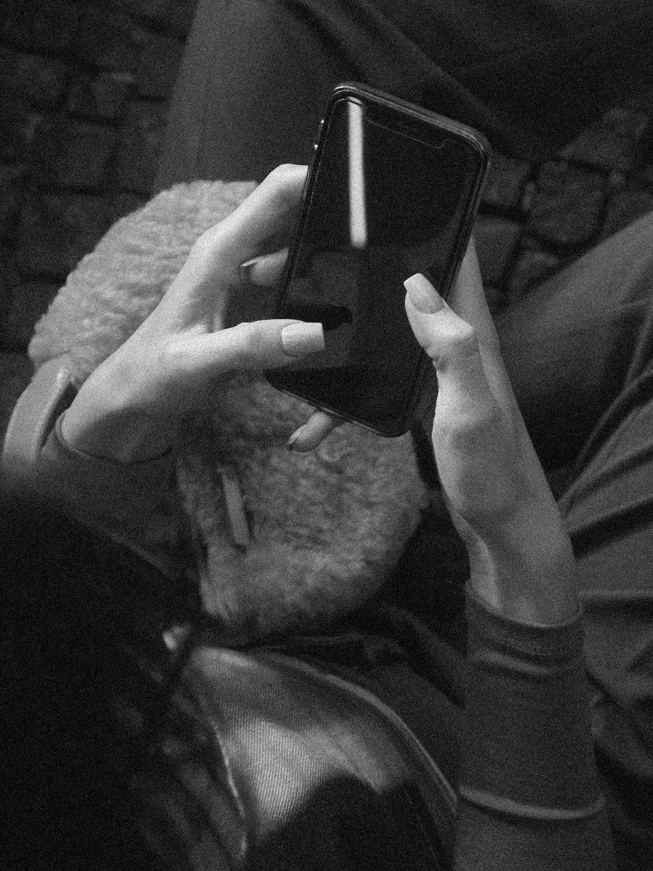A person holding a smartphone in both hands, with long manicured nails, sitting in a cozy setting with a brick wall and a blanket in the background.
