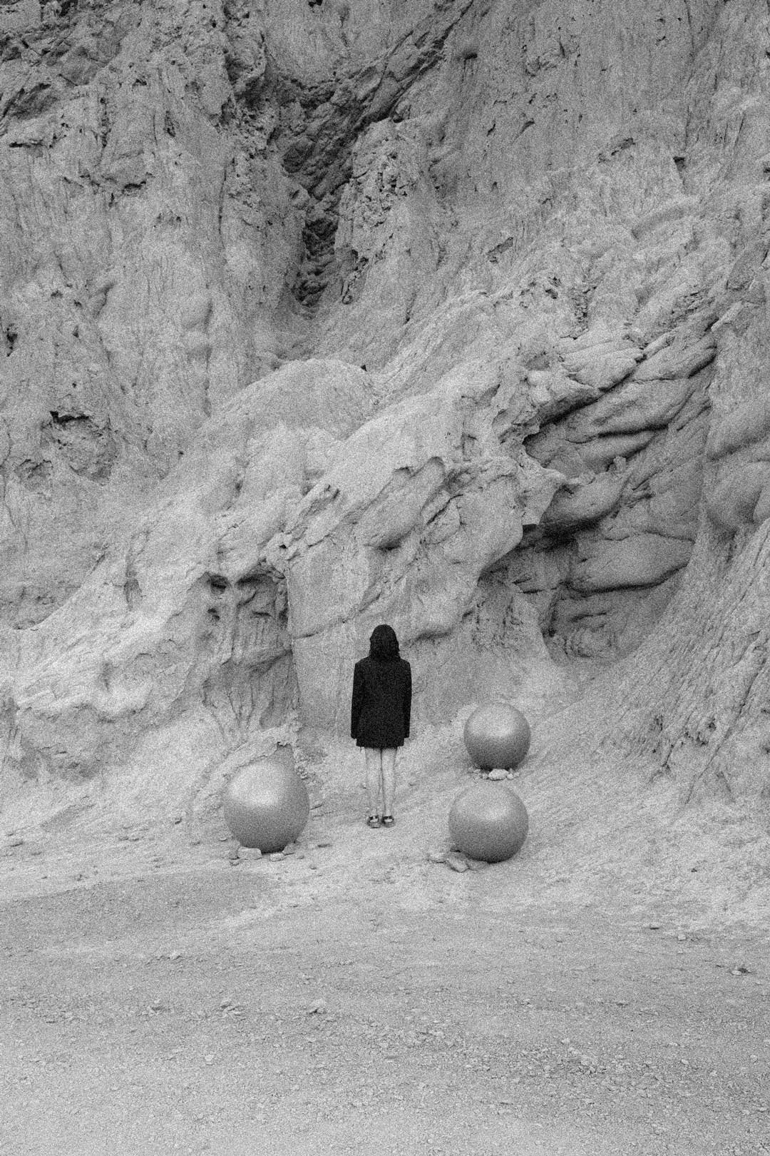 A person standing in front of a large rock formation with three large spherical objects on the ground nearby.