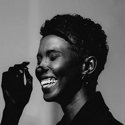 A woman with short curly hair laughing with her eyes closed, in black and white