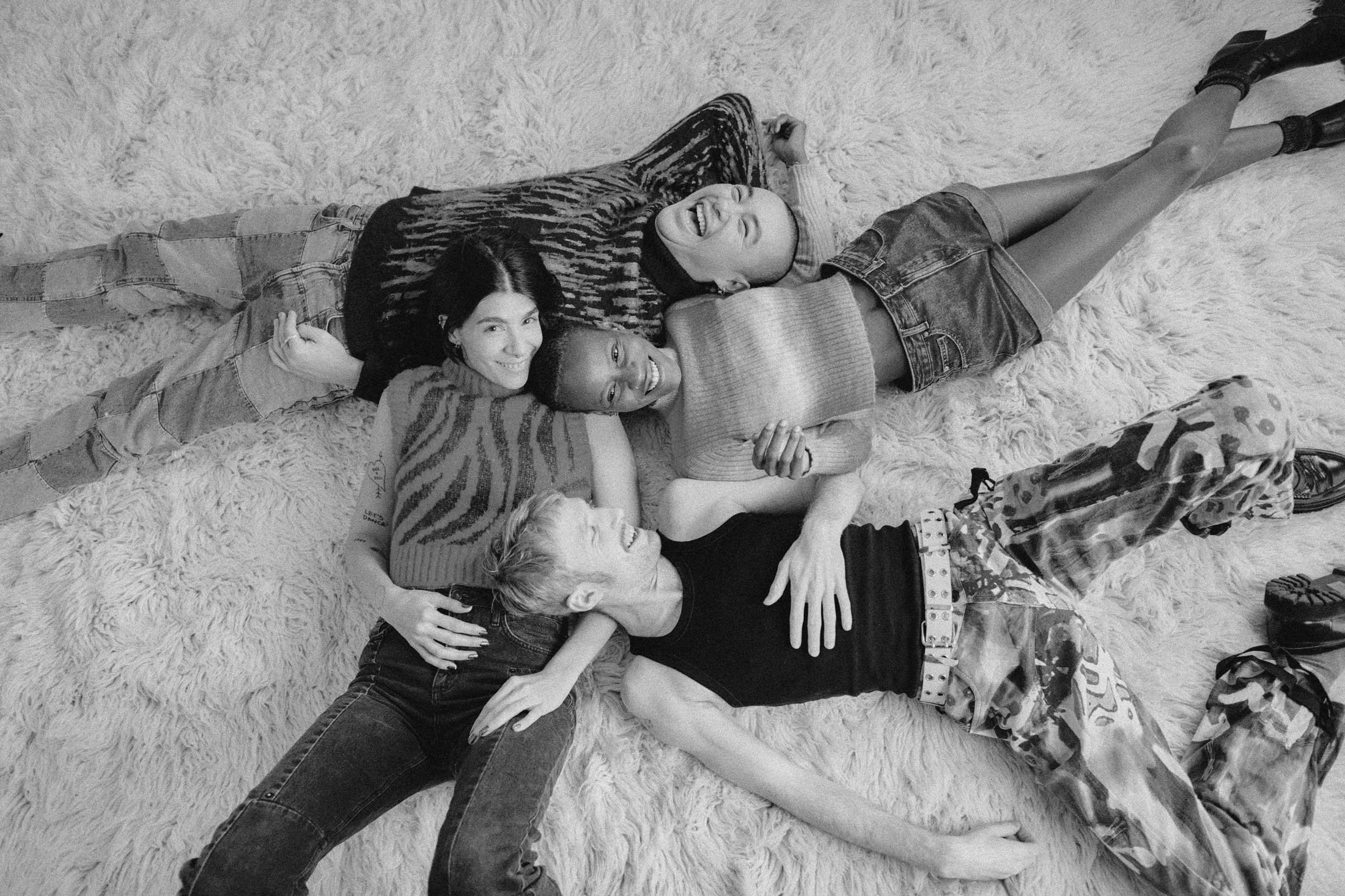 Four young women lying on a shaggy rug, smiling and hugging each other. They are wearing casual clothes and appear to be happy.