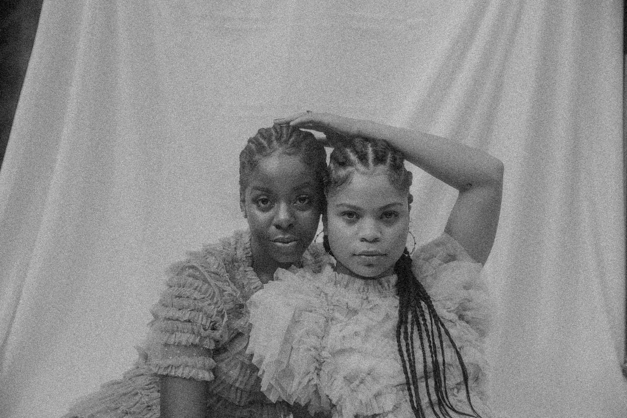 Two women with braided hair posing together in front of a curtain, one with her arm raised and resting on her head.