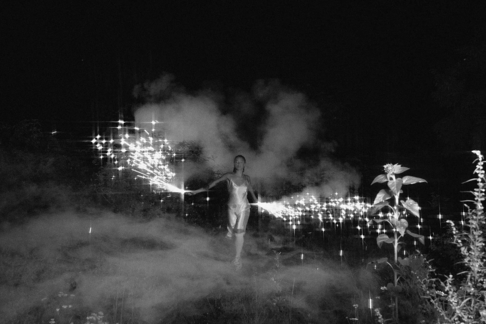 A person in a dress walking through a dark outdoor area at night holding sparklers, with smoke and trees in the background.