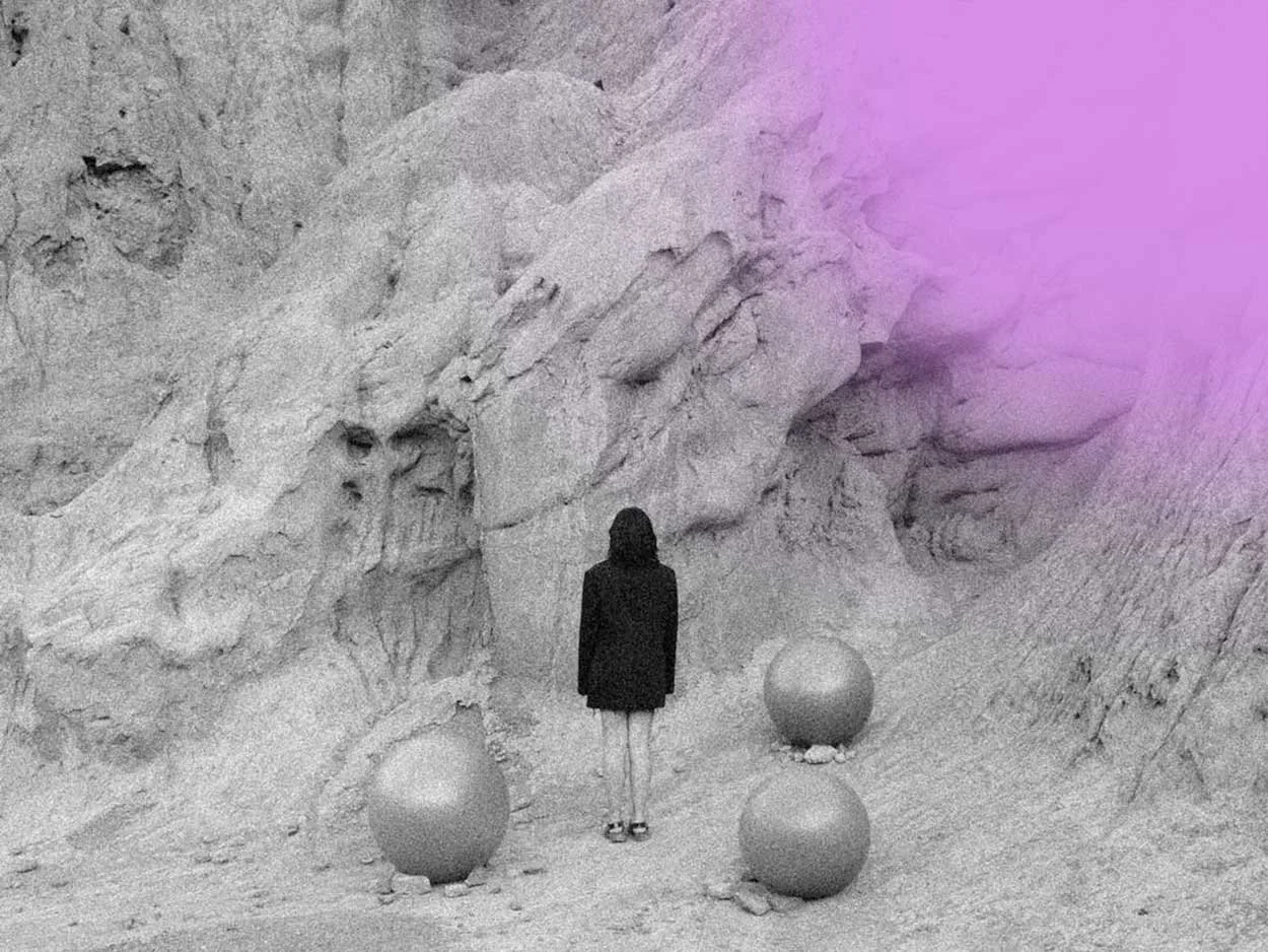 A woman in a black coat and white shoes stands in front of a rocky, desert landscape with large rocks and cliffs. There are three spherical objects on the ground near her, one on top of another. The image has a purple gradient overlay in the upper right corner.