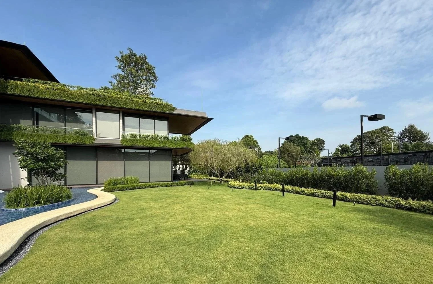 Modern house with large glass windows, surrounded by a landscaped garden with a grassy lawn, small pond, and trees under a blue sky with scattered clouds.