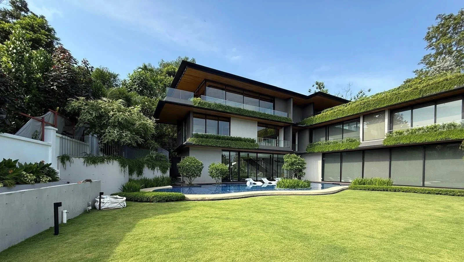Modern house with large glass windows, surrounded by a lush green lawn and trees, featuring a swimming pool with lounge chairs.