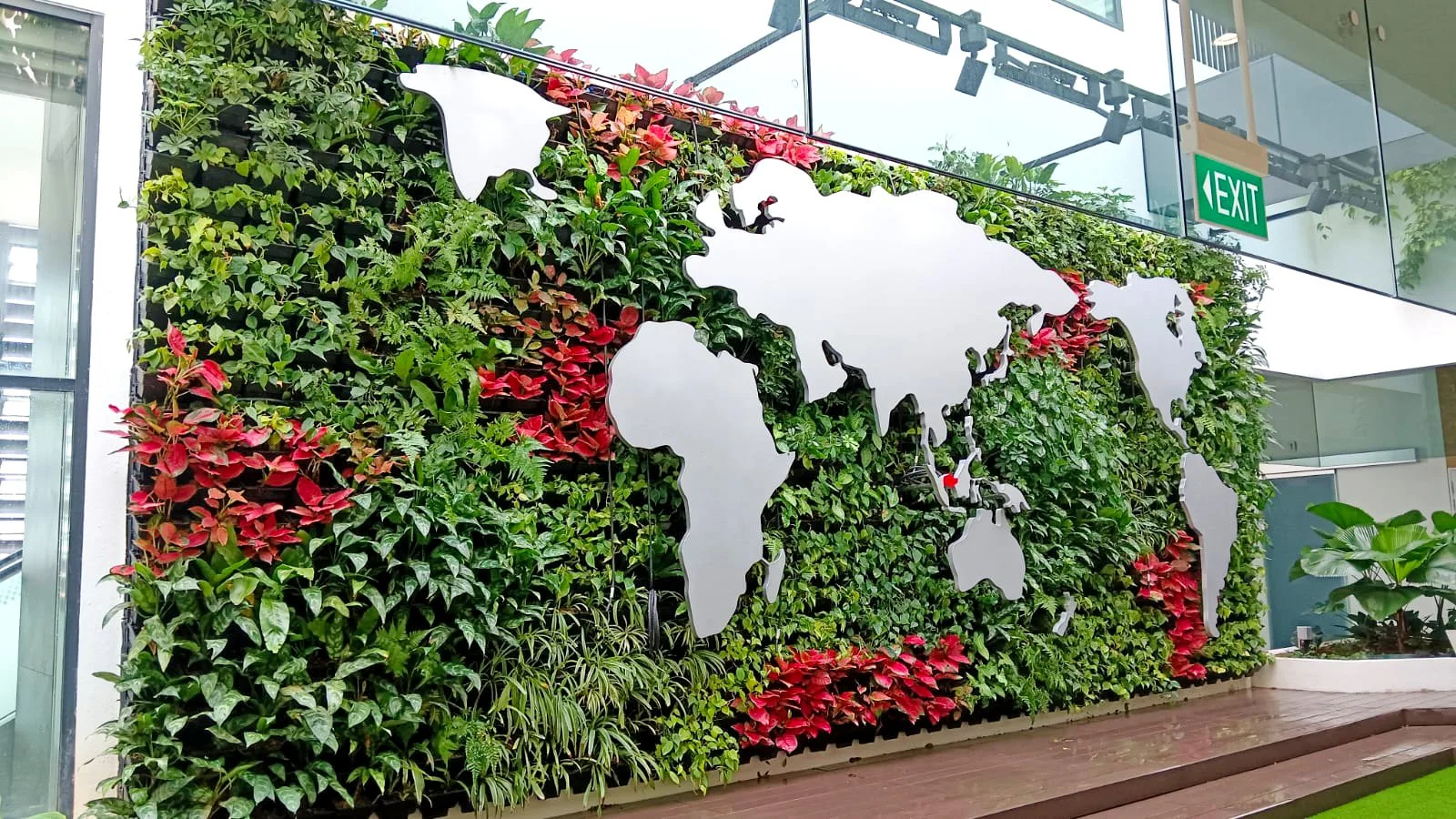 A vertical garden with various green and red plants, featuring metal cutouts of world map continents mounted on it, located inside a building near an exit sign.