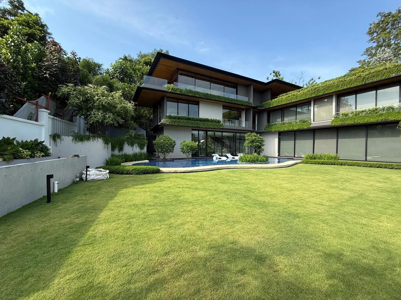 Modern house with large glass windows, surrounded by a lush green lawn and trees, featuring a swimming pool with lounge chairs.