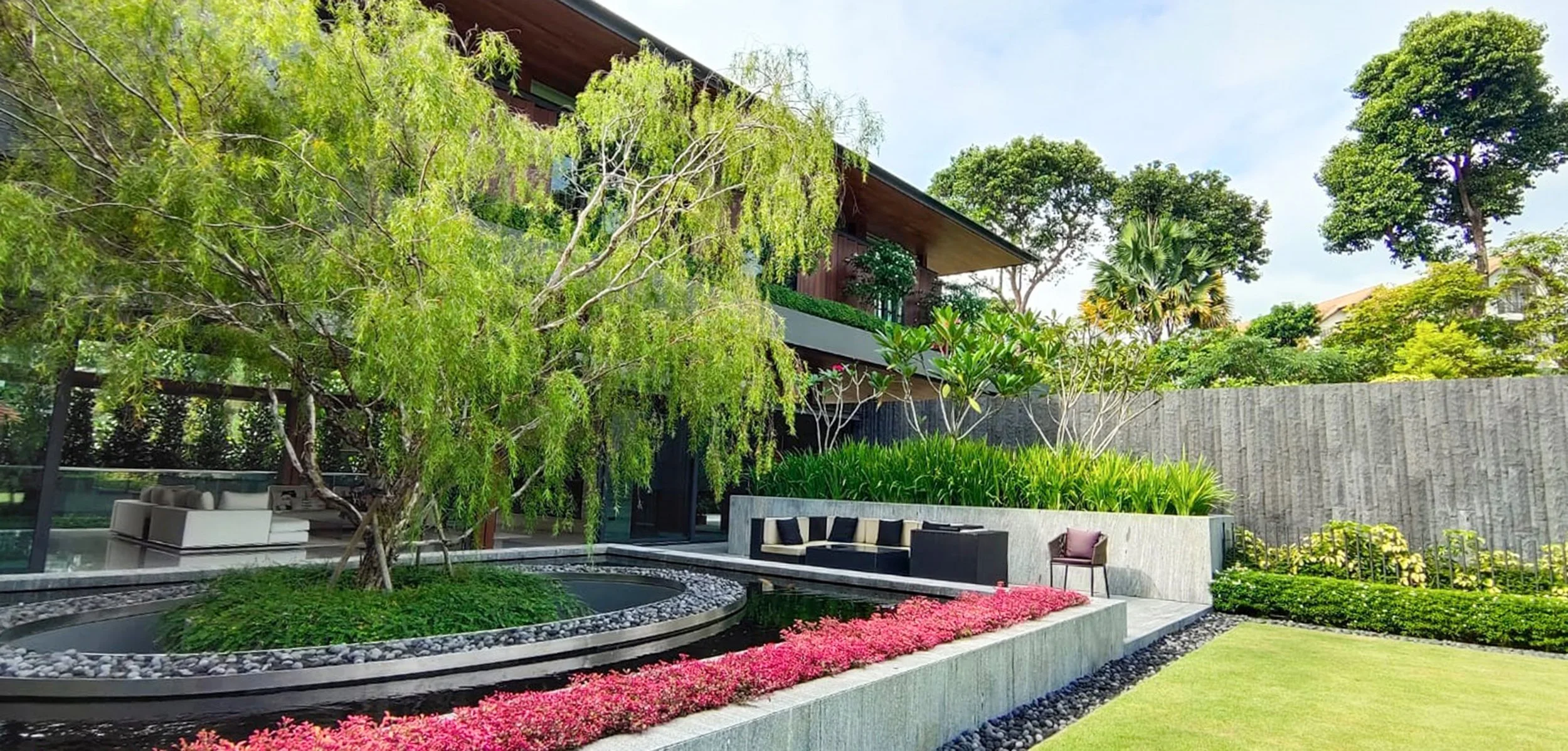 Modern backyard garden with lush greenery, a curved water feature surrounded by decorative rocks, vibrant pink flowers, and outdoor seating areas with a contemporary house in the background.