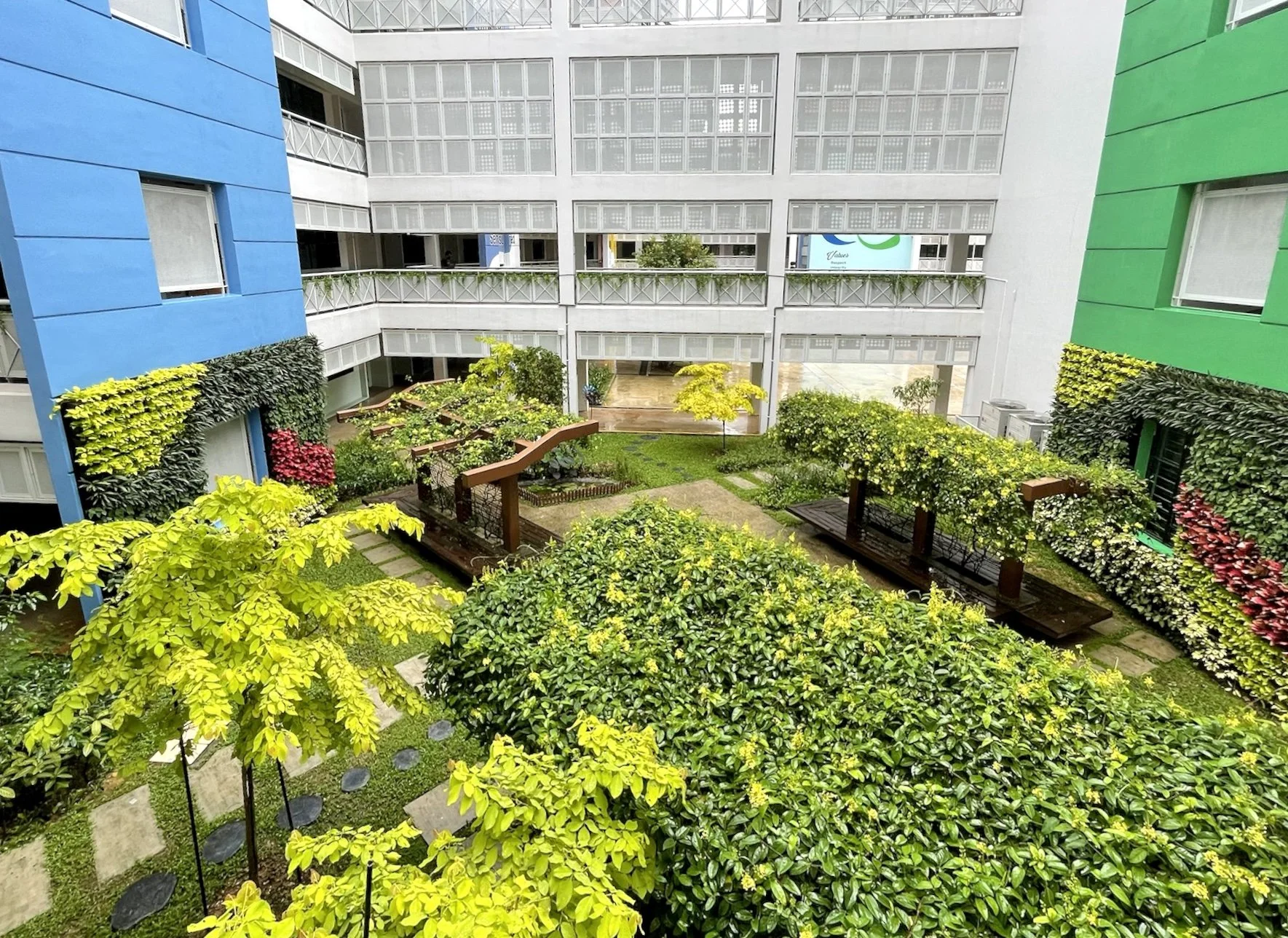 A spacious courtyard garden framed by colorful apartment buildings painted blue and green. The garden features lush green trees, shrubs, and decorative plants, with pathways and wooden benches for seating.