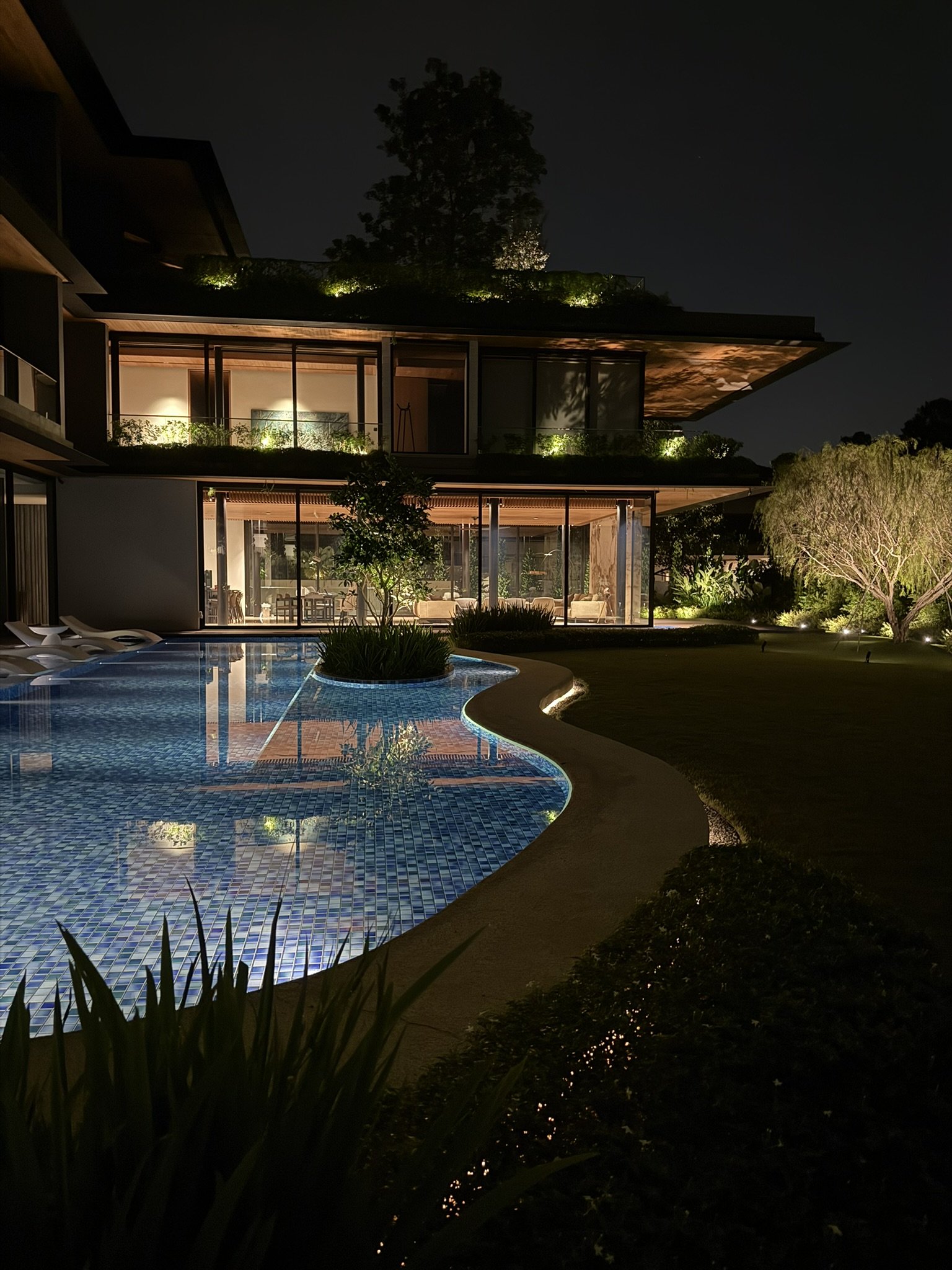 Nighttime view of a modern house with large glass windows, illuminated from inside. In the foreground, there is a uniquely shaped swimming pool with blue tile lining, surrounded by a garden with small lights. The house has multiple floors with balcon