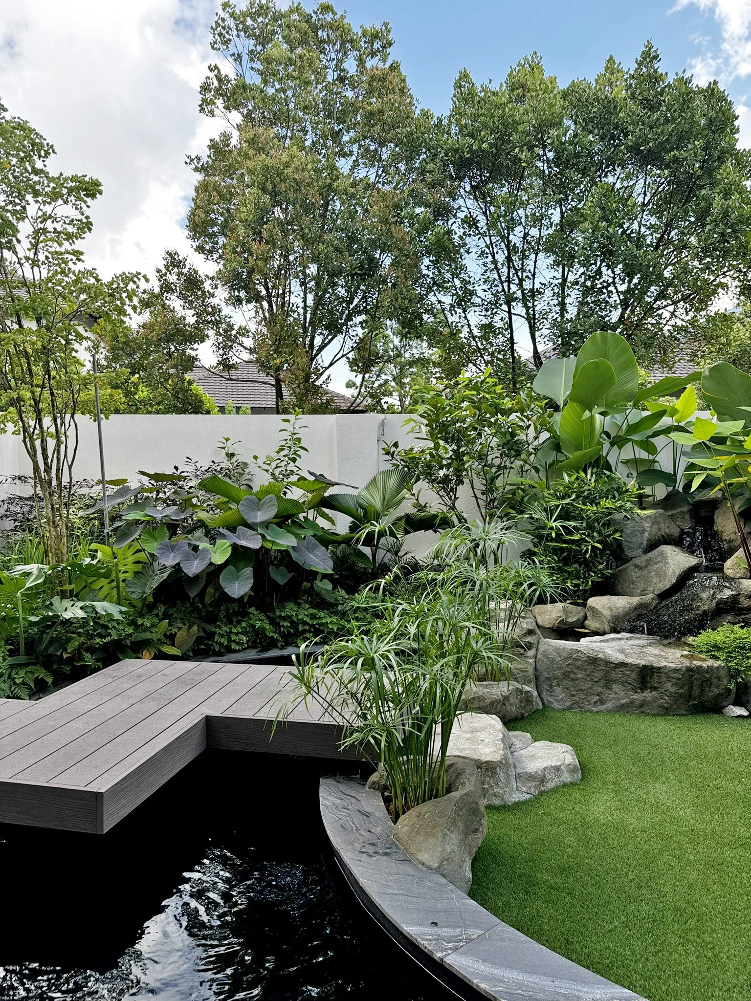 Backyard garden with lush green plants, large rocks, and a small waterfall. There is a wooden platform over a pond, a white fence, and trees in the background under a partly cloudy sky.