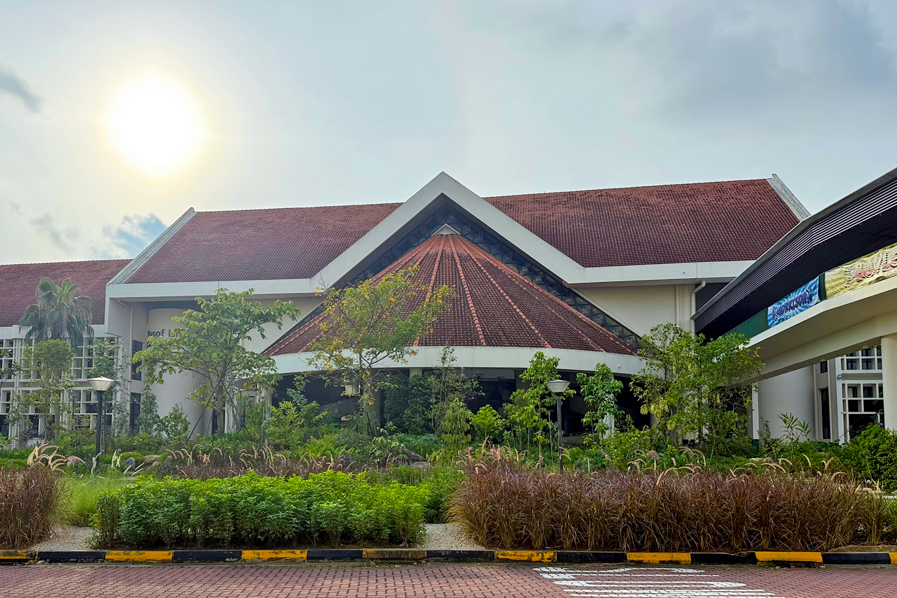 A large building with a pyramid-shaped roof, surrounded by a landscaped garden with trees, shrubs, and ornamental grasses. The sky is partly cloudy with the sun shining.