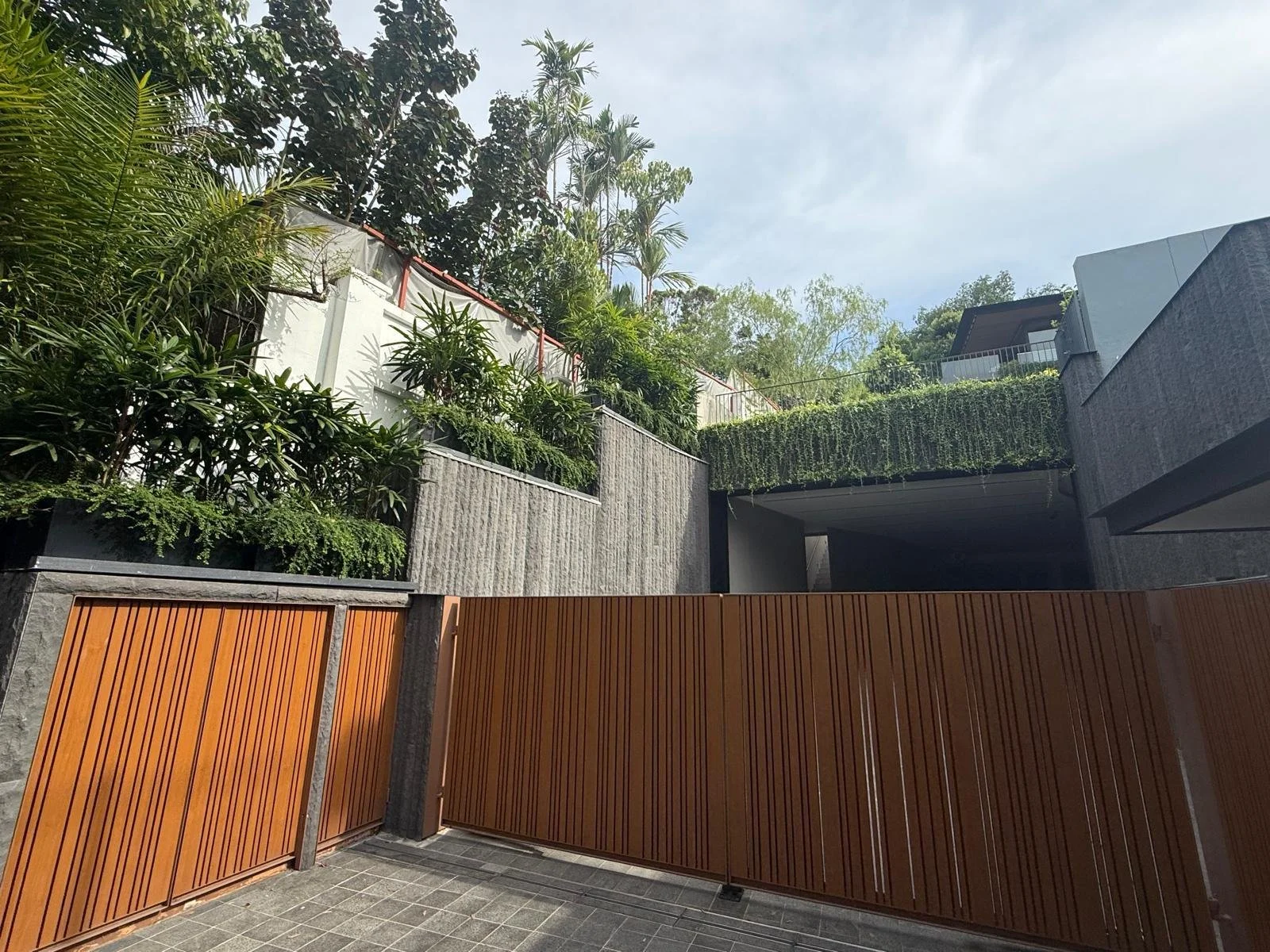Modern residential building with gray and black exterior walls, a wooden gate, and lush greenery including trees and bushes in a landscaped yard.