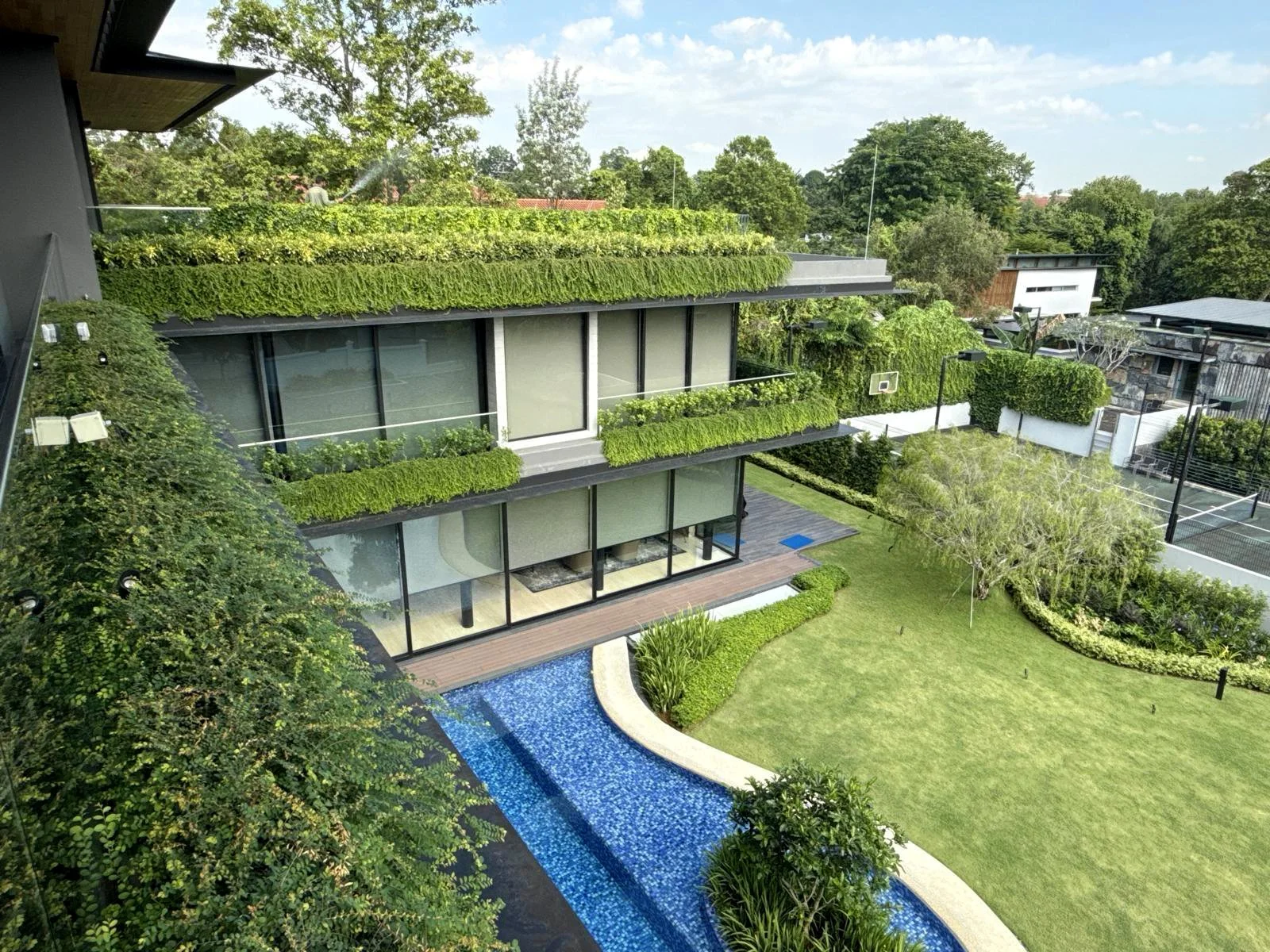 Modern multi-story house with green rooftop gardens, large glass windows, a swimming pool, and a well-maintained grassy yard surrounded by trees and shrubbery.