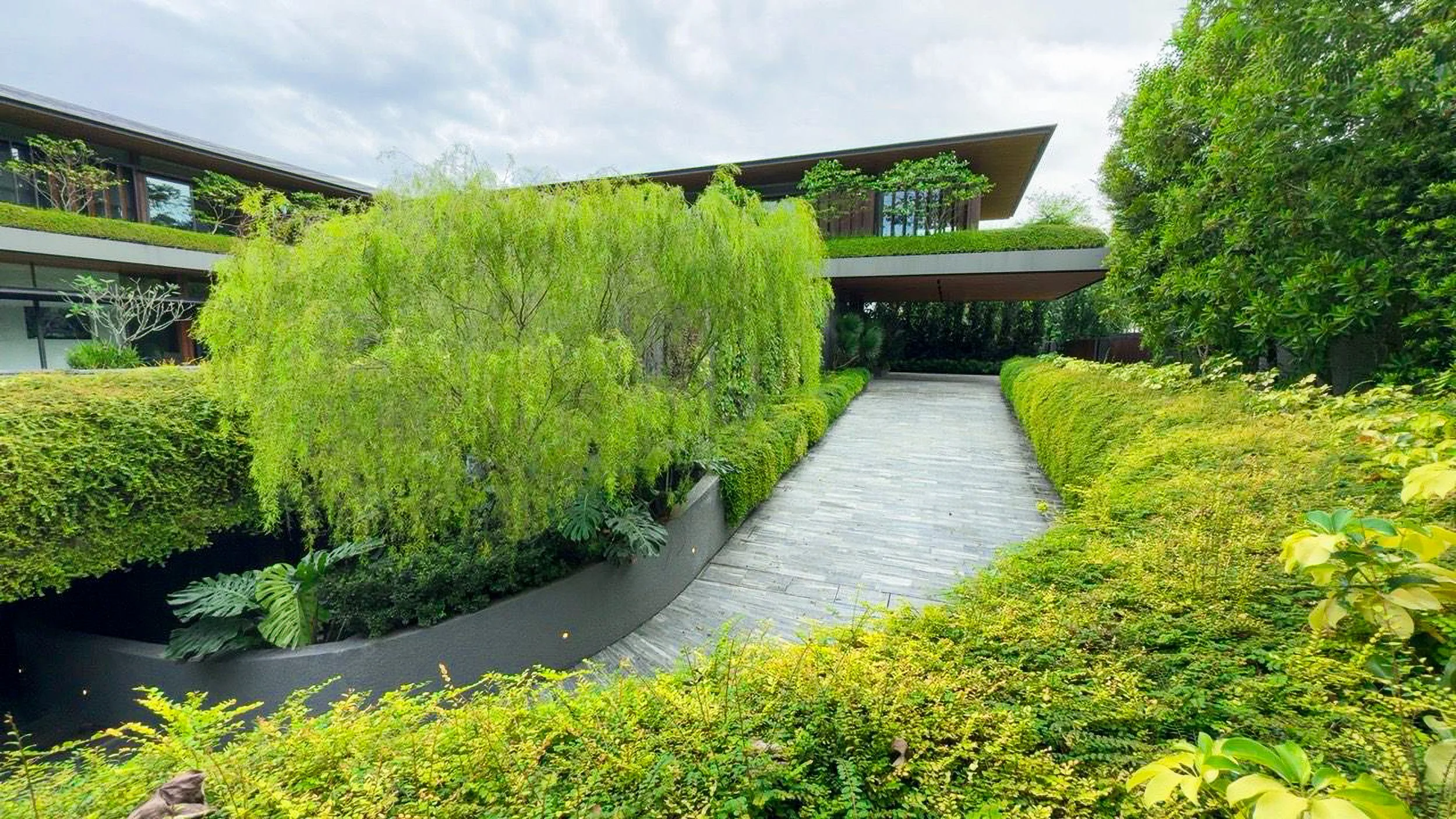 A modern building with an overhanging roof surrounded by lush green shrubs and trees, with a paved pathway leading through the garden.