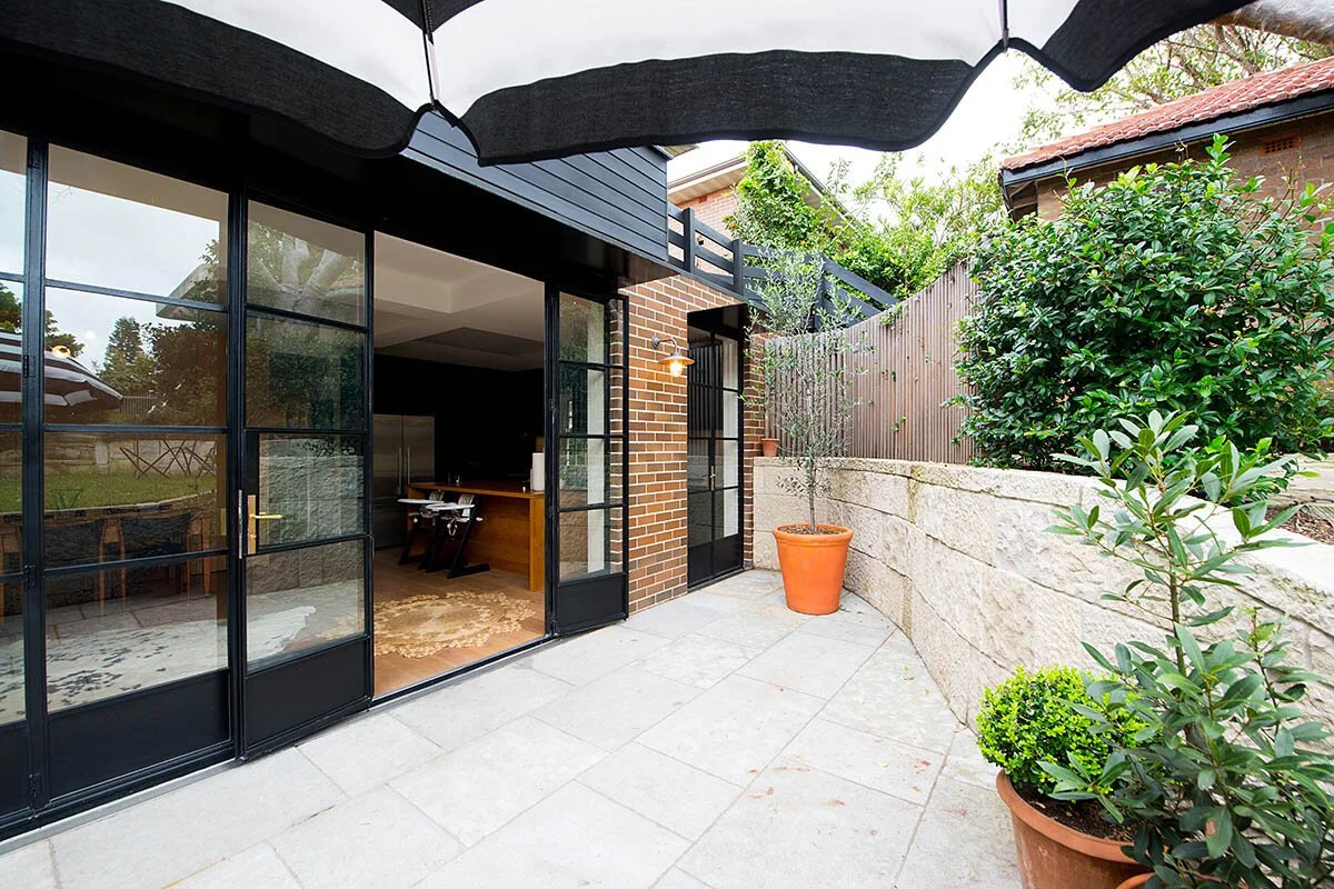 View of a modern house patio with large glass sliding doors, potted plants, and a high brick and wood fence, with a balcony above and a black umbrella partially visible at the top.