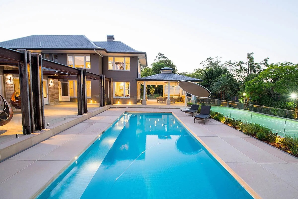 Modern backyard with swimming pool, outdoor lounge area, and tennis court, surrounded by trees and landscape lighting.