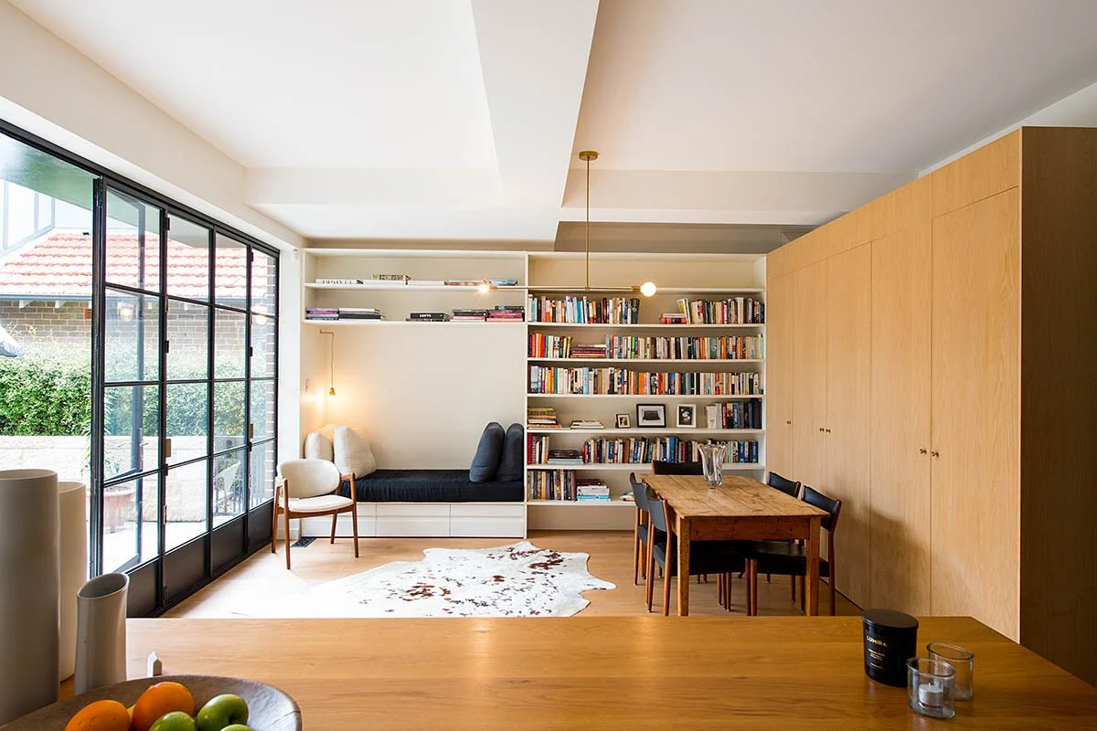A cozy living room with a large window, a built-in couch with cushions, a bookshelf filled with books, a wooden dining table with chairs, and minimal modern decor.
