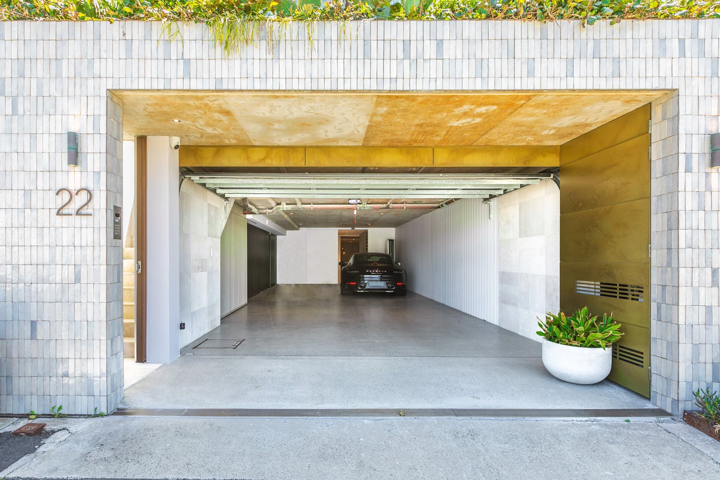 Modern residential garage with a black car parked inside, stone and metal exterior, house number 22, potted plant on the right, and beige tile wall with greenery at the top.