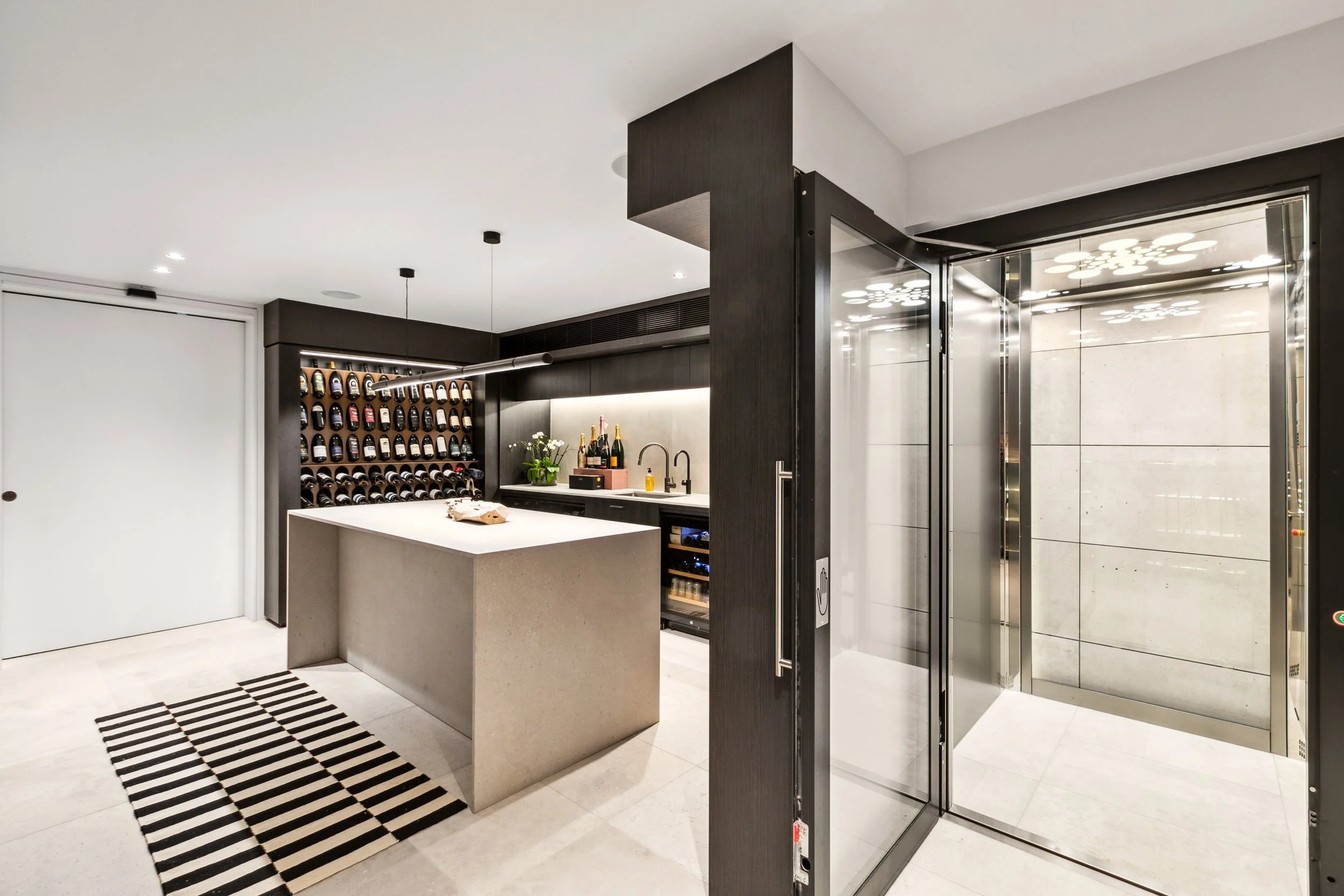 Modern wine cellar with wine racks, a kitchen island, and an elevator door on a tiled floor.