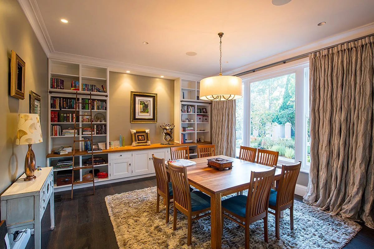 A dining room with a wooden table and six matching chairs, a bookshelf and cabinet against a wall, a large window with curtains, a chandelier hanging above the table, and a rug on the dark hardwood floor.