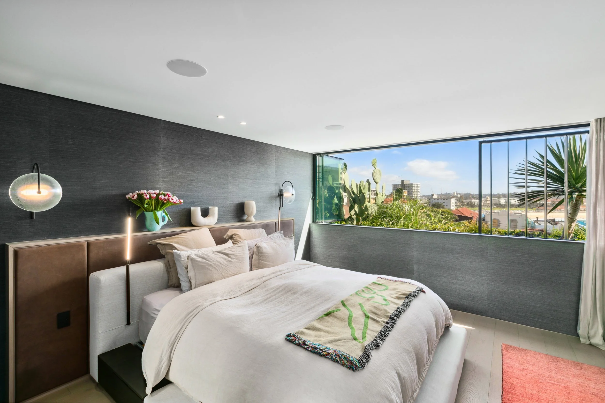 Modern bedroom with large window view of city and cacti, with bed, pillows, and decorative wall art.