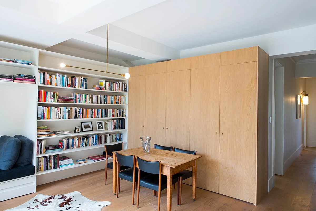 A cozy living space with a white bookshelf filled with books, a small wooden dining table with four black chairs, and a wooden cabinet or closet to the right. The room has wooden floors and modern lighting fixtures.
