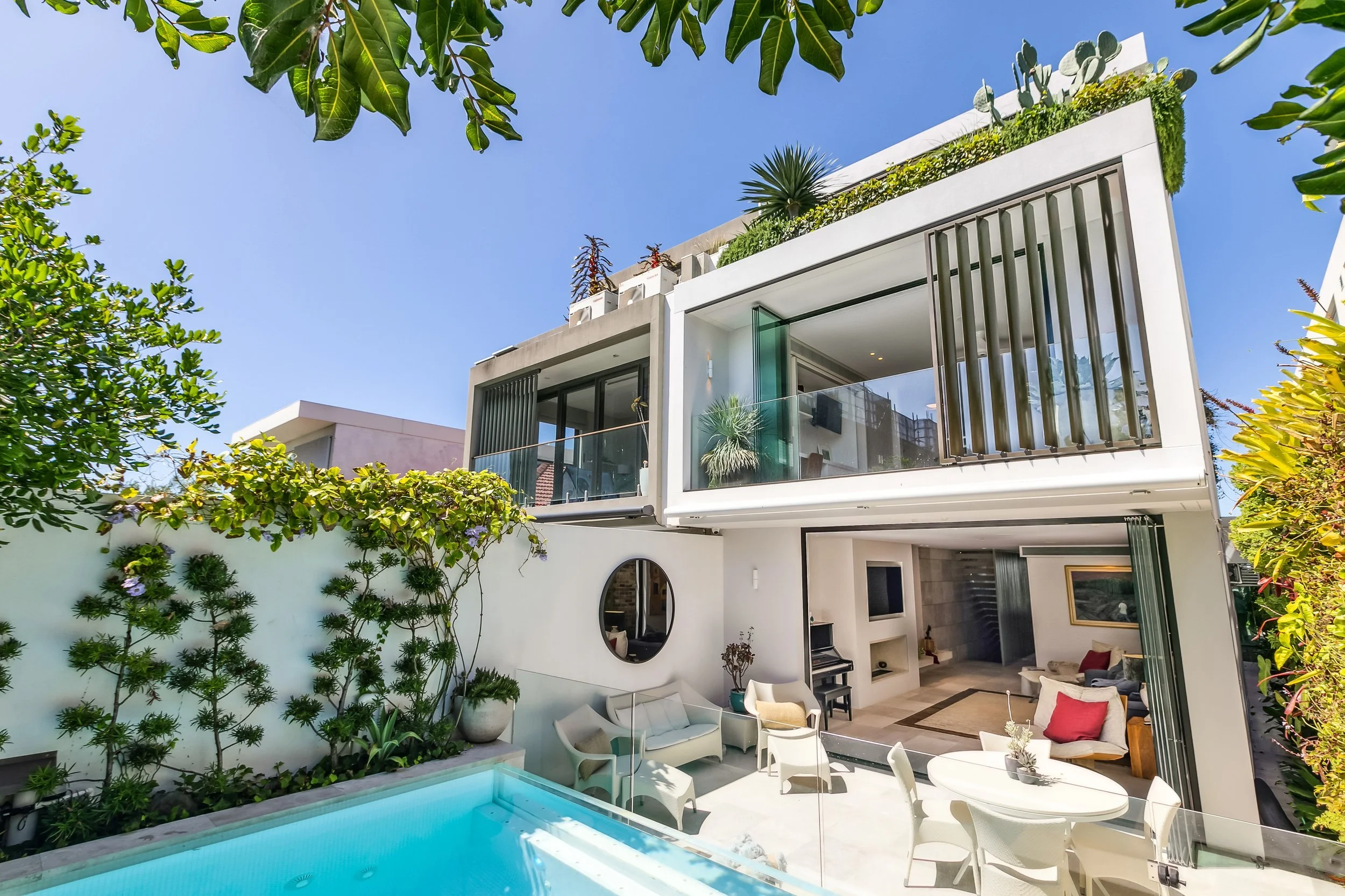 Modern multi-story house with large glass windows and balcony, surrounded by greenery, with an outdoor pool and seating area in the yard.