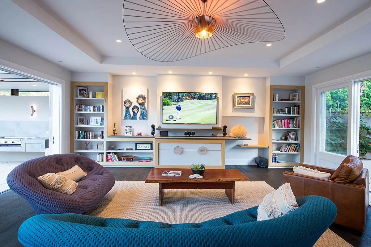 Living room with colorful curved sofas, a wooden coffee table, a wall-mounted TV, built-in bookshelves, and large windows showing greenery outside.
