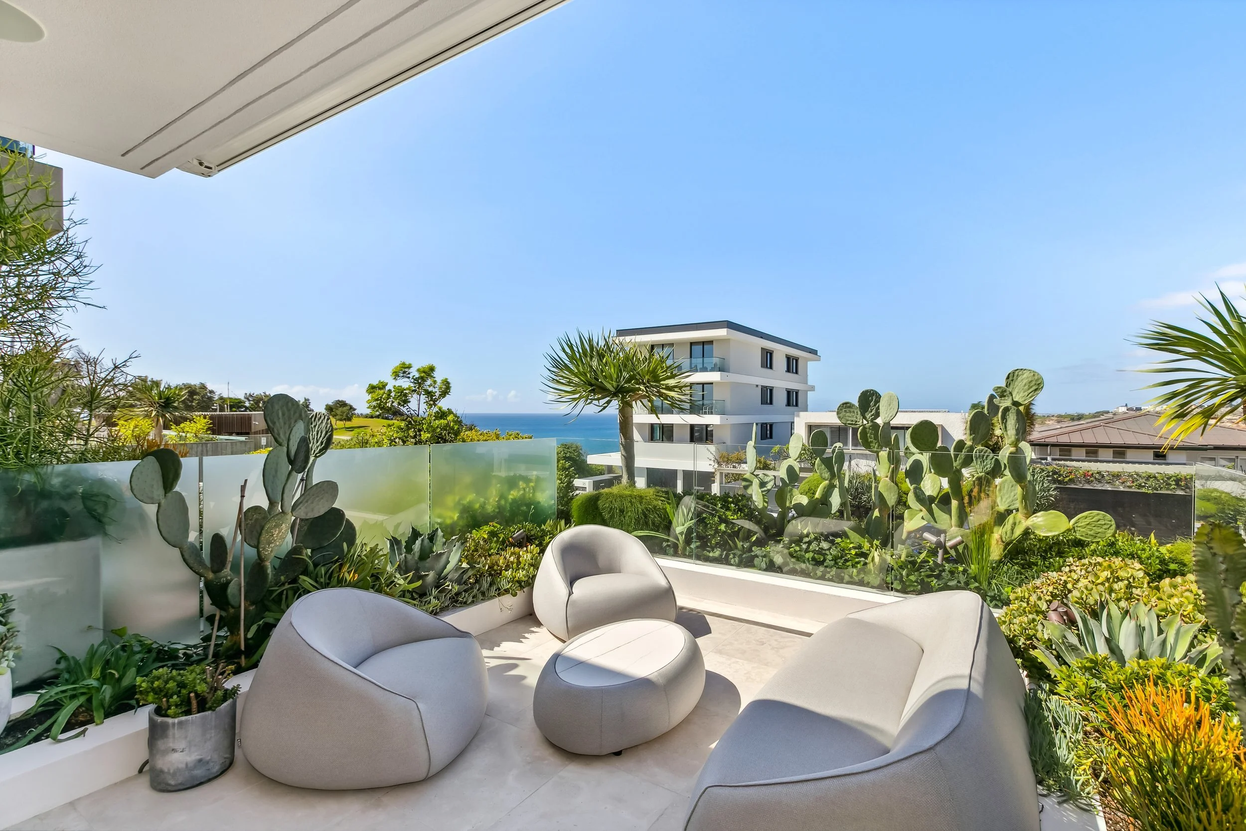 Balcony with modern white furniture and potted plants, overlooking trees, a building, and the ocean in the distance on a sunny day.