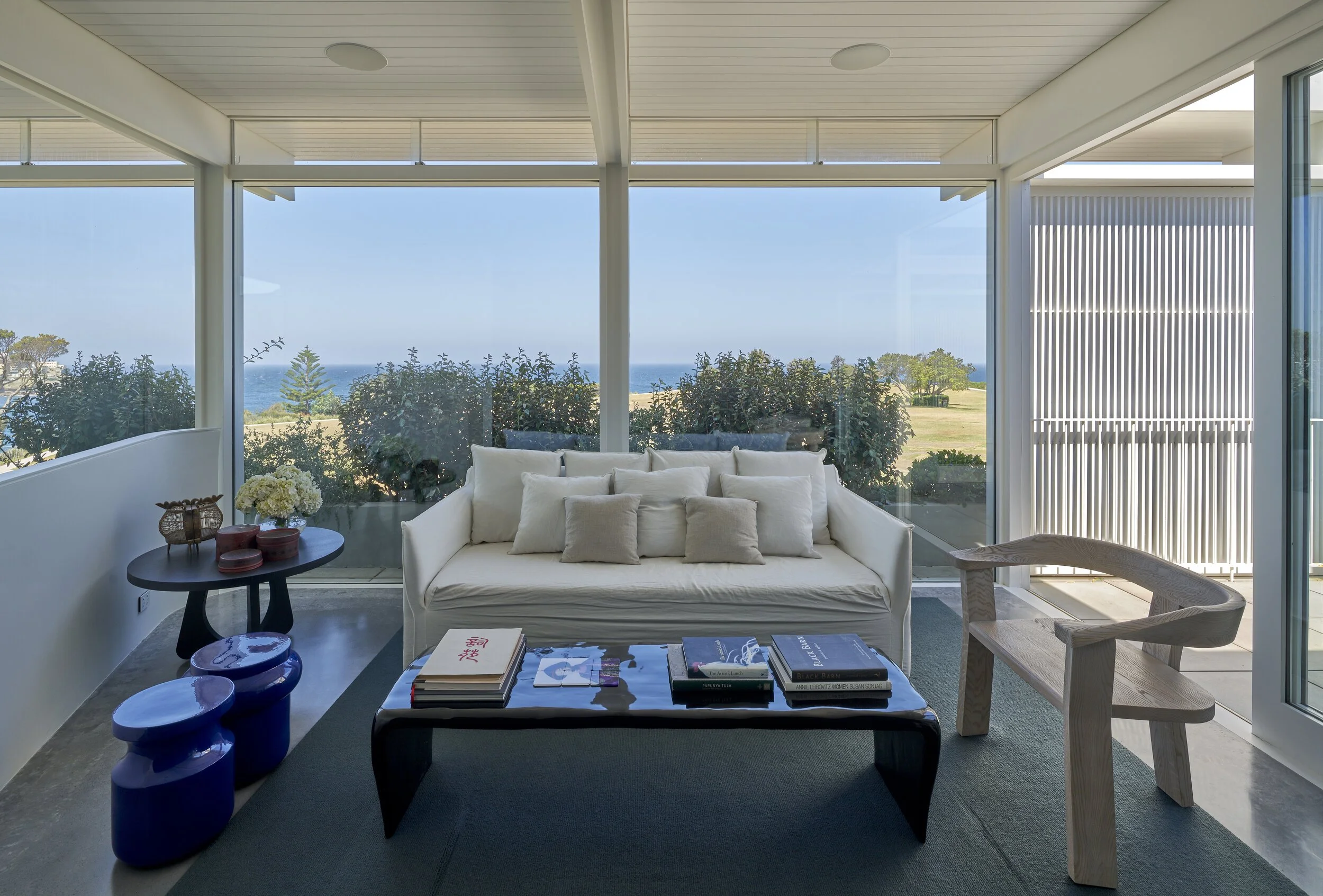Living room with large windows overlooking ocean, white sofa with cushions, black coffee table with books, side table with flowers, wooden chair, and decorative blue stools