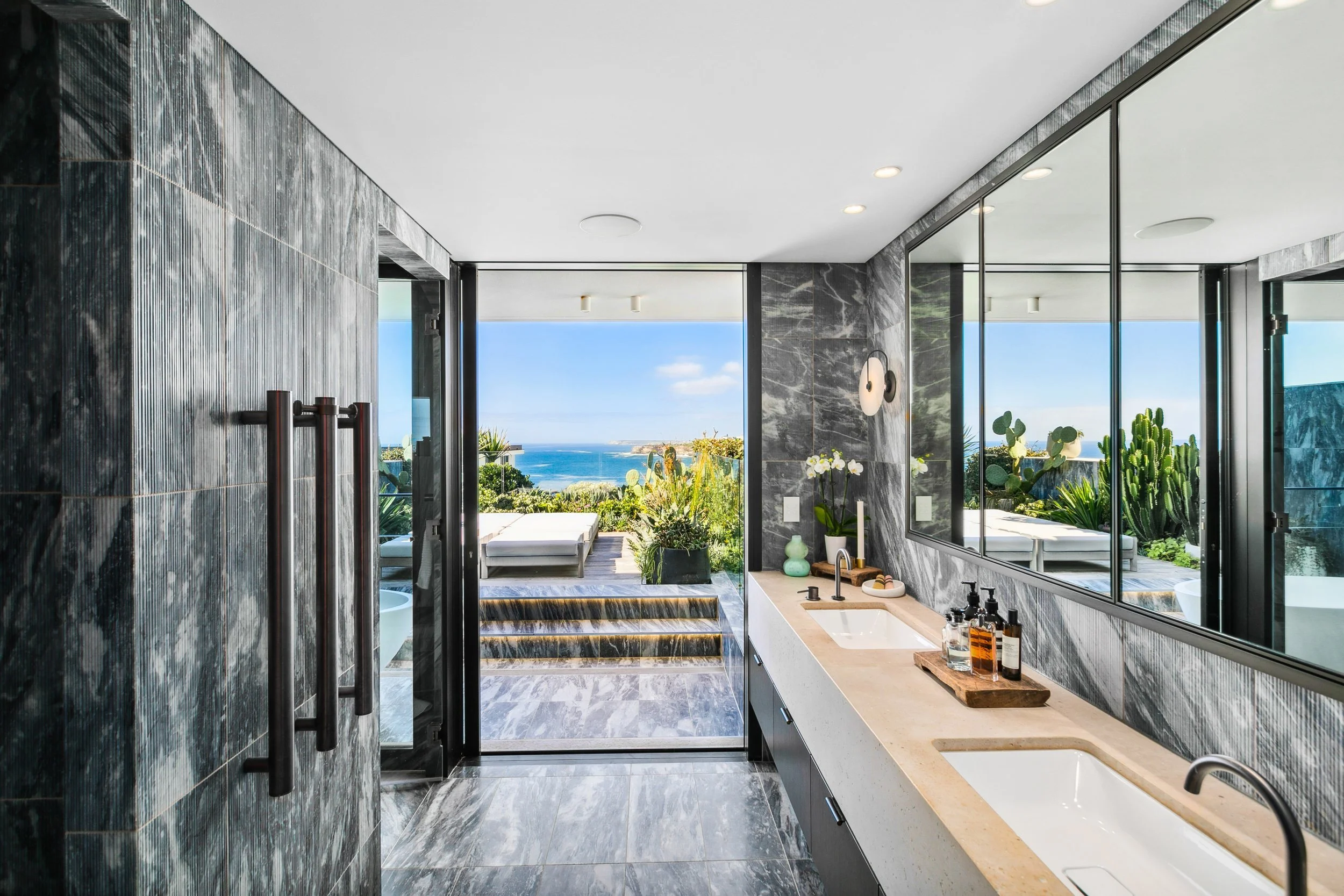 Modern bathroom with marble walls and floors, a double sink vanity, large mirror, and a door leading to an outdoor patio with a view of the ocean, plants, and outdoor furniture.