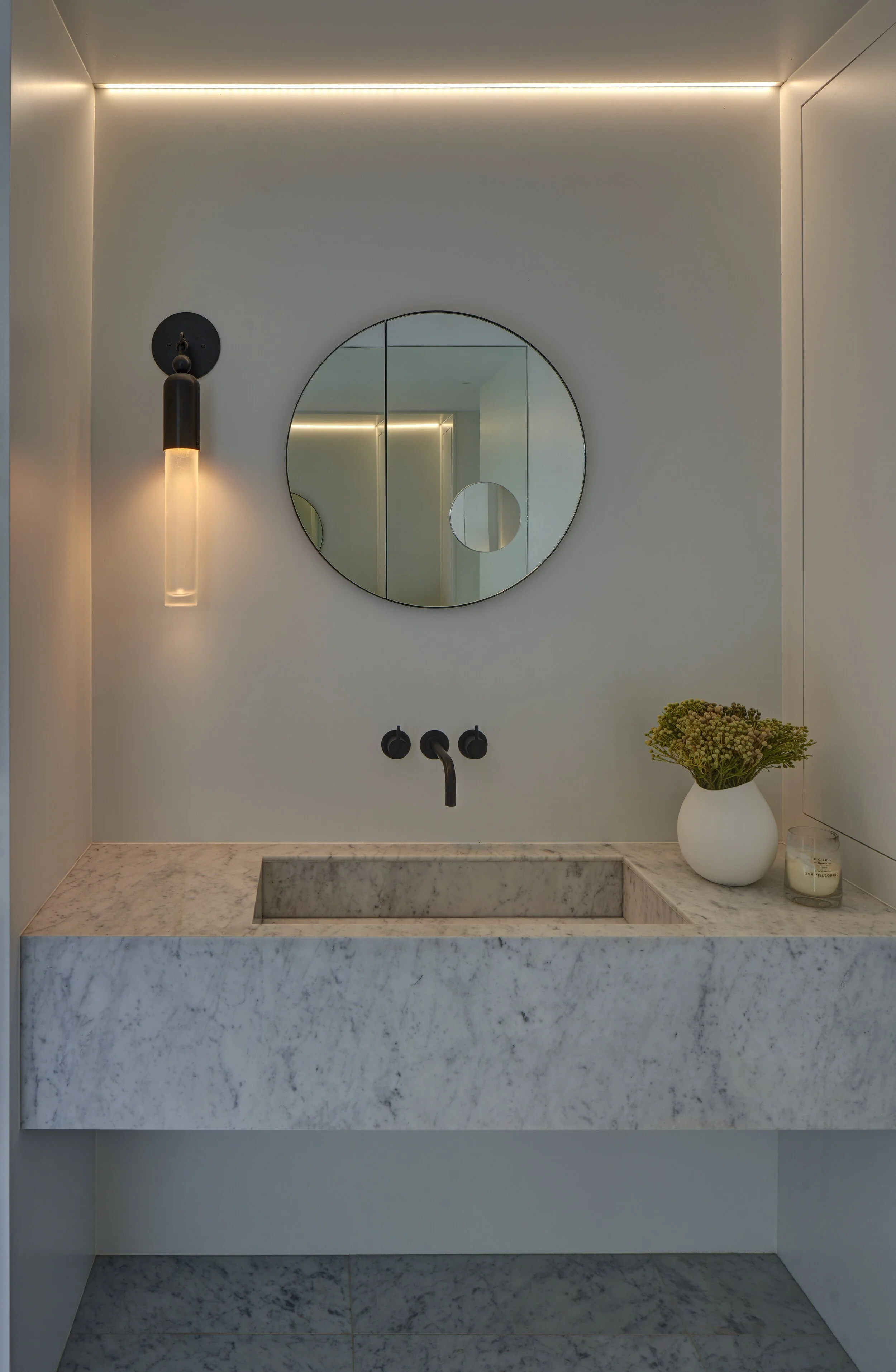 Modern bathroom vanity with a white marble countertop, built-in sink, black faucet, round mirror, black wall-mounted light, a vase with green flowers, and a candle in a glass holder.