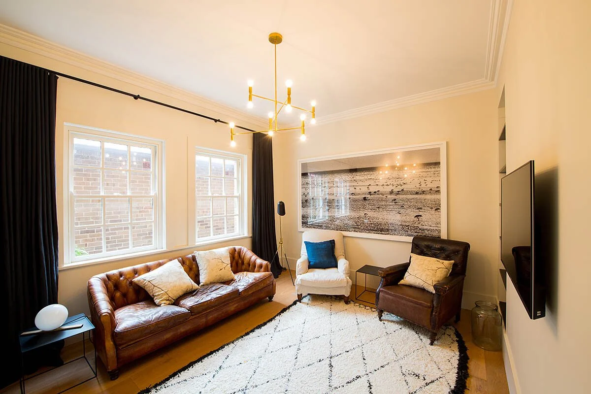 Living room with a leather sofa, a white armchair, a brown armchair, a large framed black-and-white photograph on the wall, a flat-screen TV on the wall, a white shag rug, and windows with dark curtains.