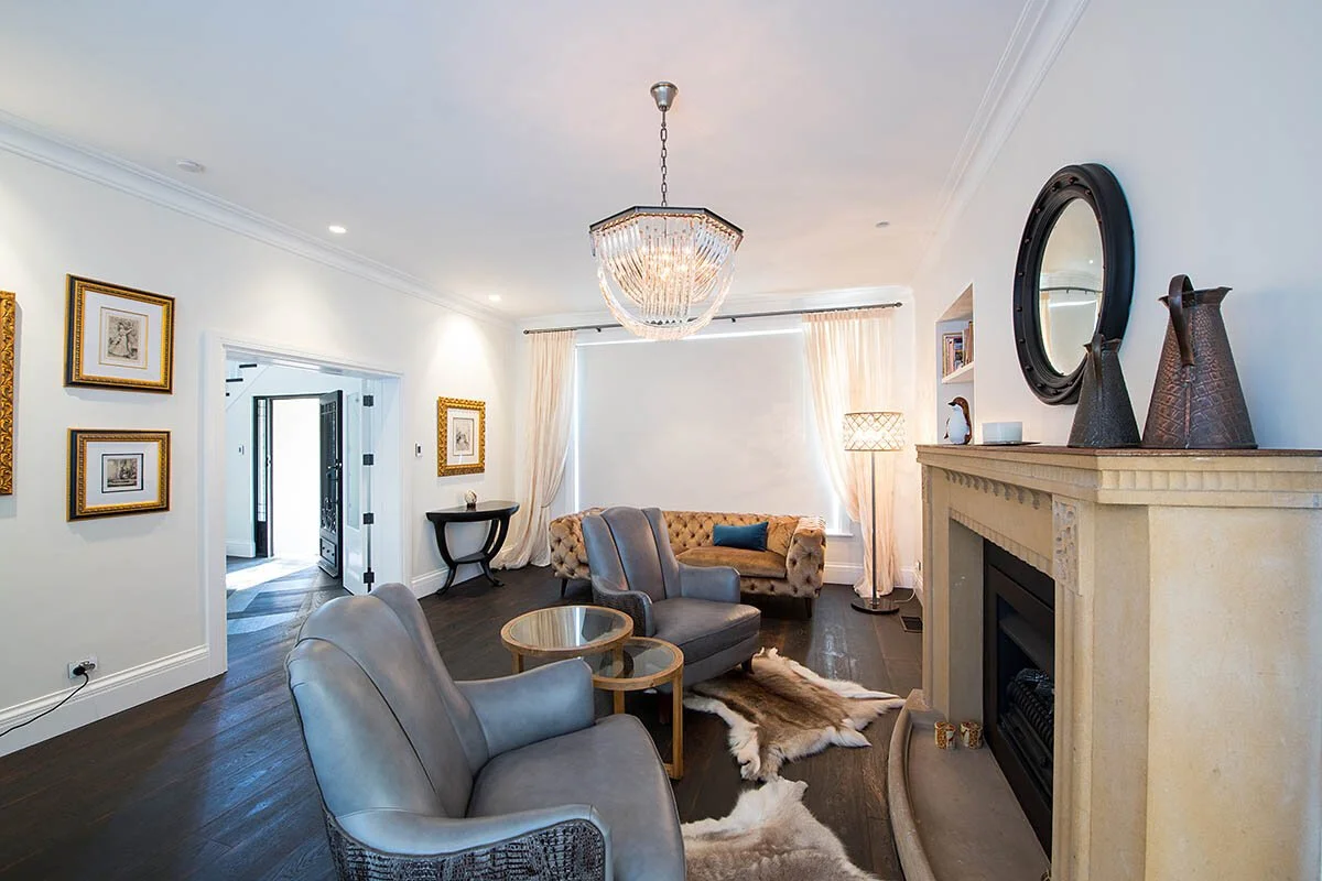 Living room with a chandelier, cream walls, dark wooden floor, and a fireplace. There are two gray armchairs, a beige tufted sofa with a blue pillow, a round side table, a cowhide rug, and a standing lamp. Framed artwork hangs on the walls, and there is a mirror above the fireplace with decorative vases.